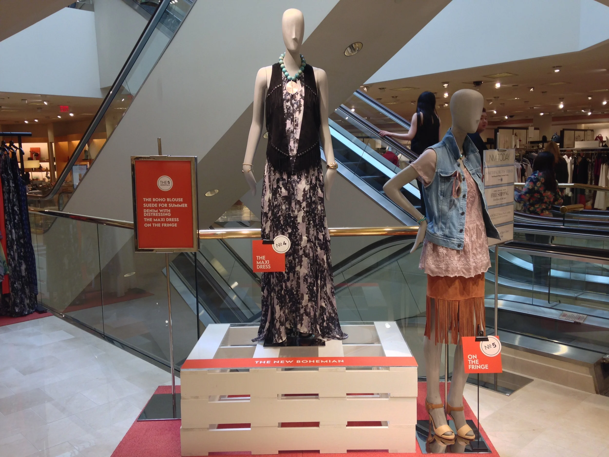 Mannequins dressed in fashionable summer outfits displayed in a retail store with escalators in the background. One mannequin wears a sleeveless maxi dress with a black and white pattern, accessorized with a turquoise necklace and bracelets, while an