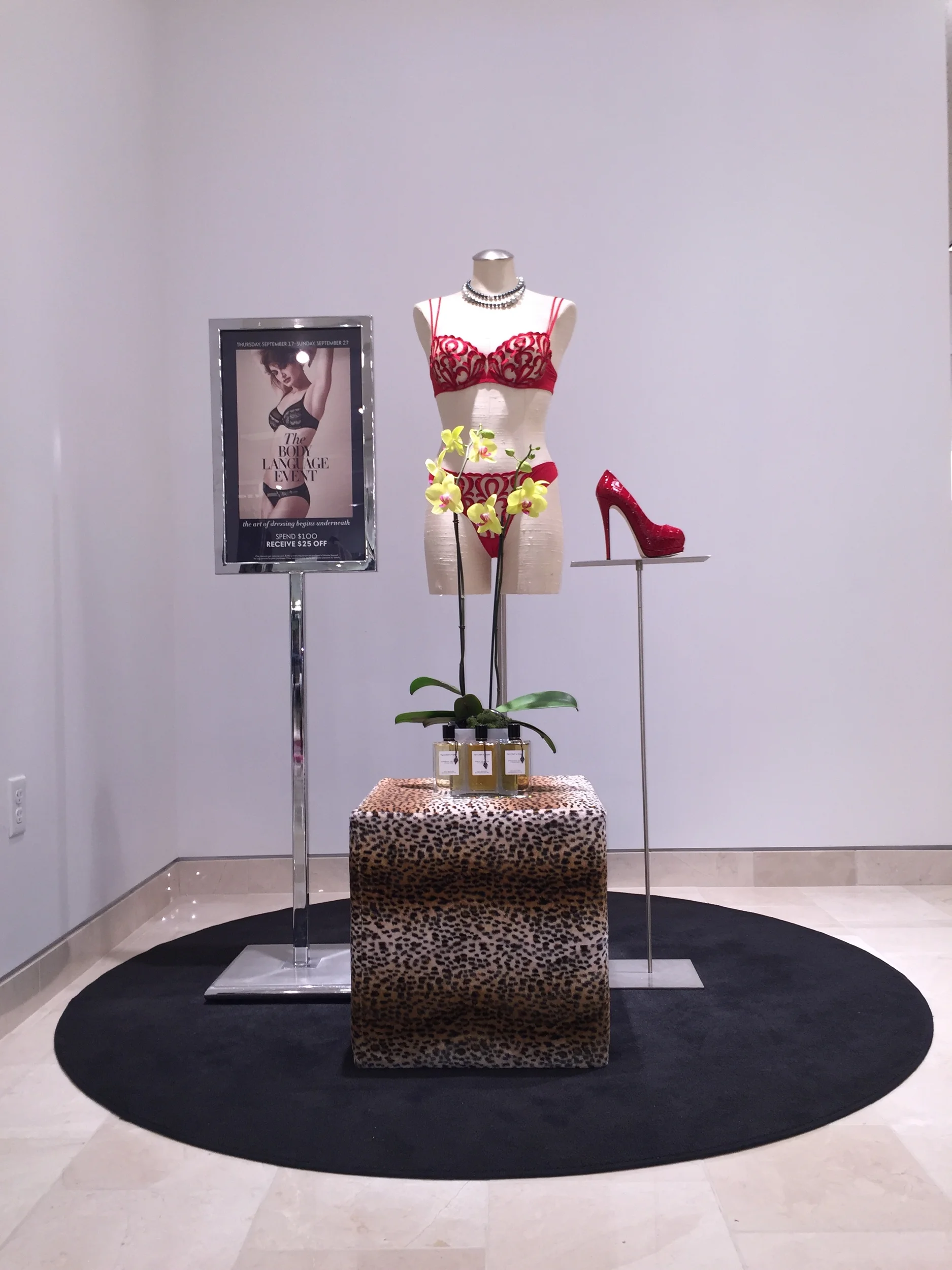 Display of a mannequin dressed in red lace lingerie with necklaces, a red high heel shoe, a potted yellow orchid, and a leopard print table covering on a black oval rug