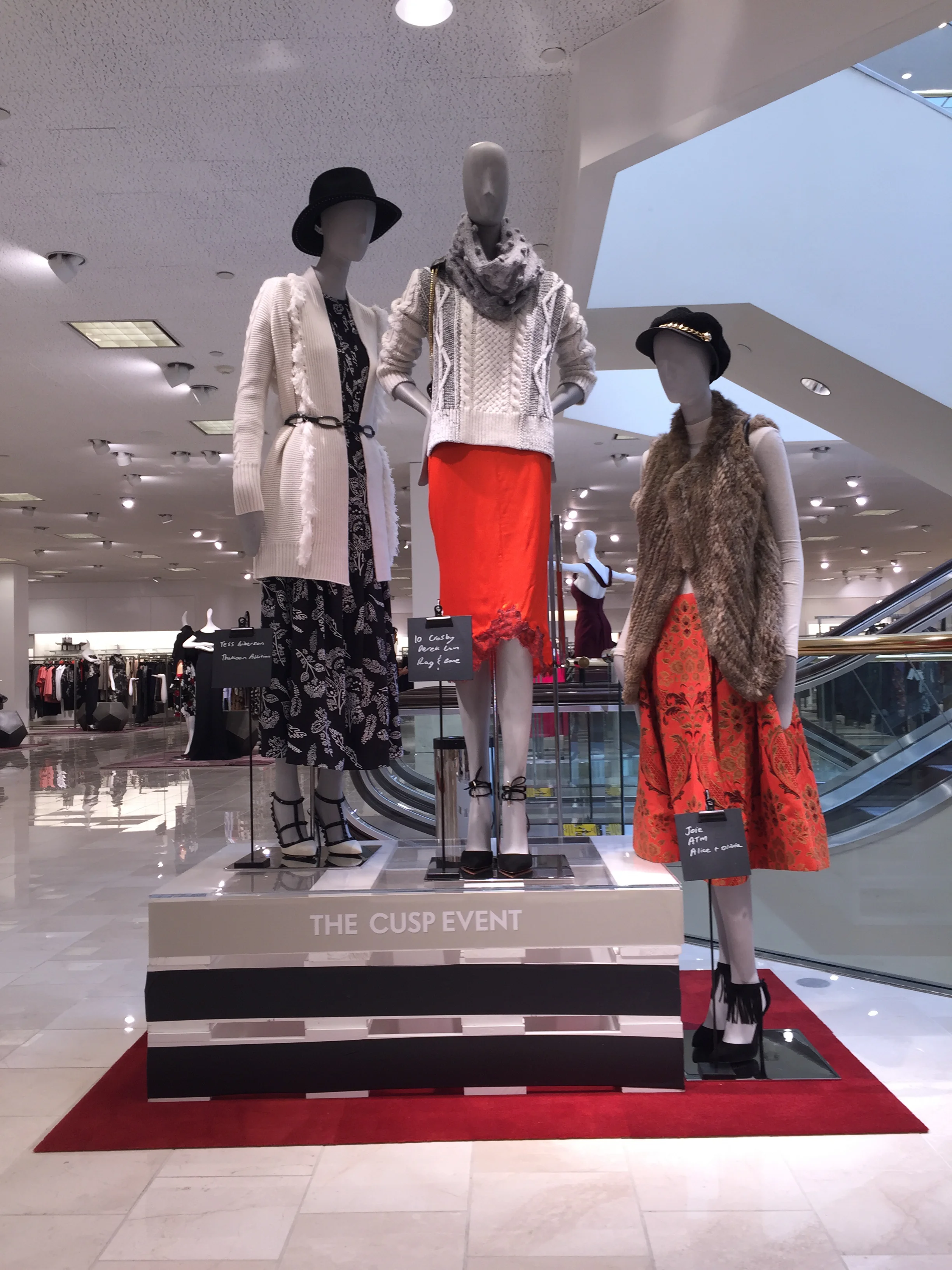 Display of three mannequins dressed in fashionable clothing at a retail event called 'The Cusp Event' inside a shopping mall. The mannequins showcase stylish outfits with accessories, with a background of shopping racks and bright interior lighting.