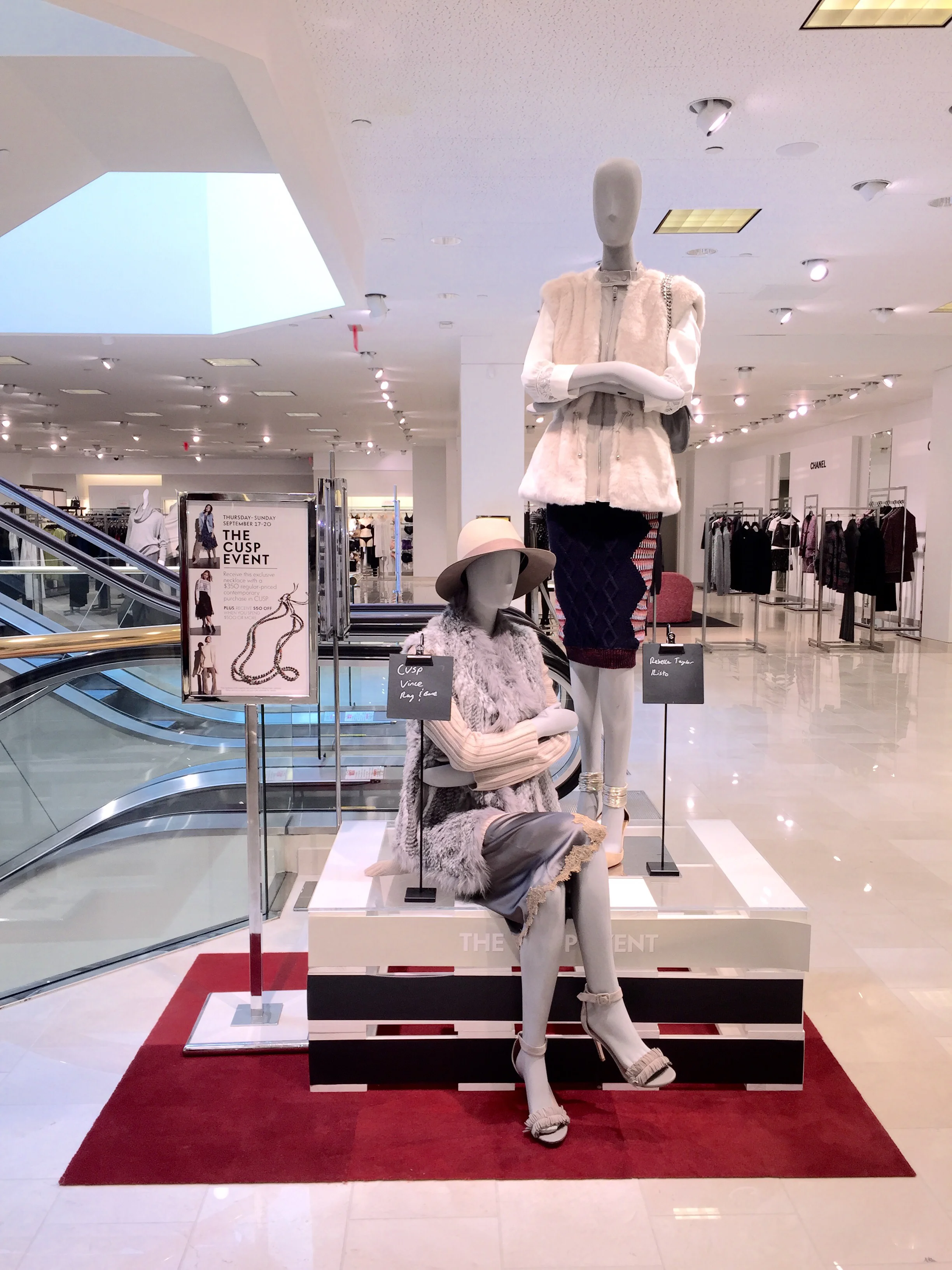 Mannequin dressed in white and fur clothing on display in a shopping mall, with a red carpet and promotional sign for a jewelry event nearby.