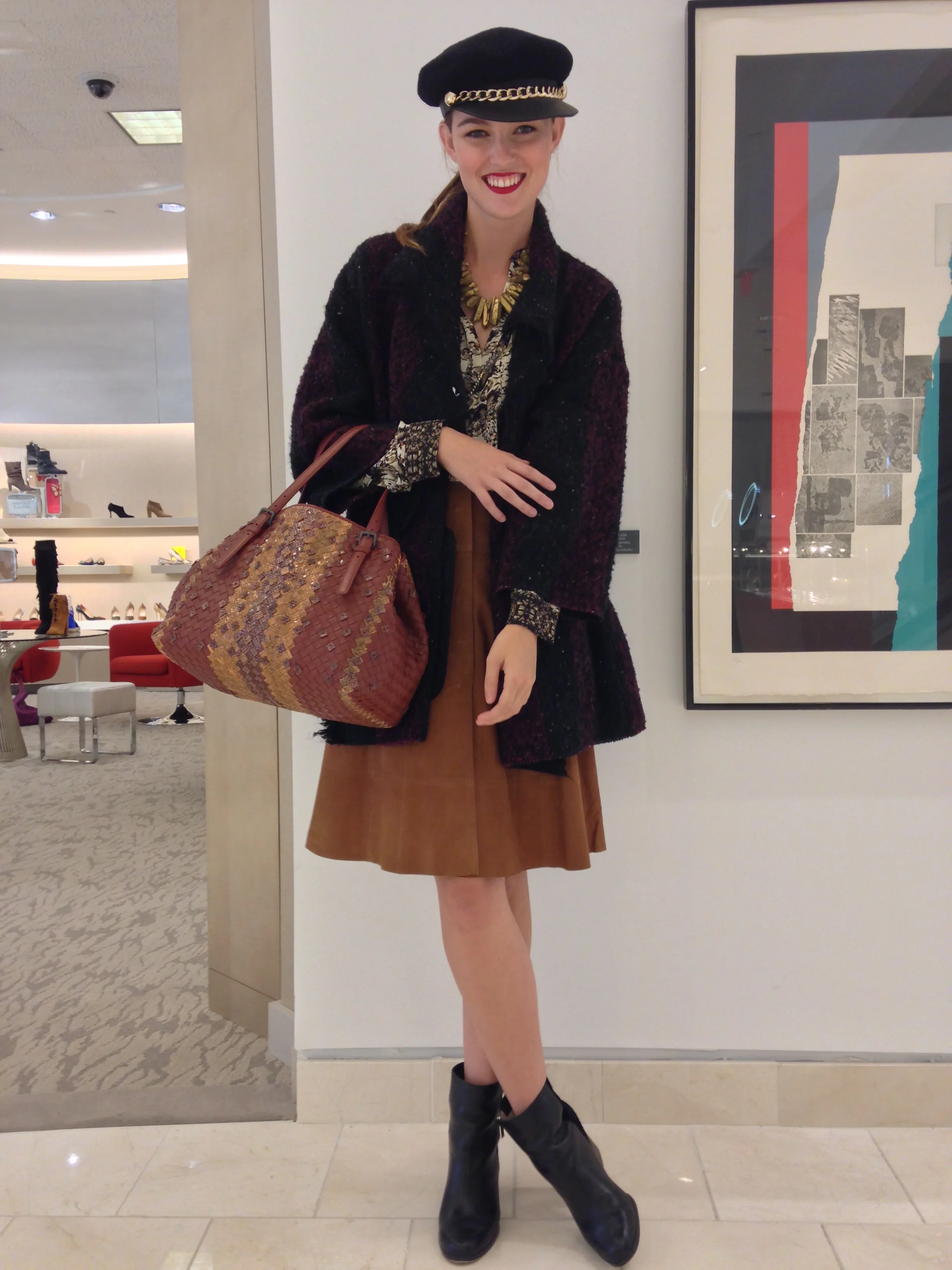 A woman standing indoors, wearing a black hat with a gold chain, red lipstick, a patterned top, a brown skirt, a dark textured coat, and knee-high black boots, holding a large woven handbag.