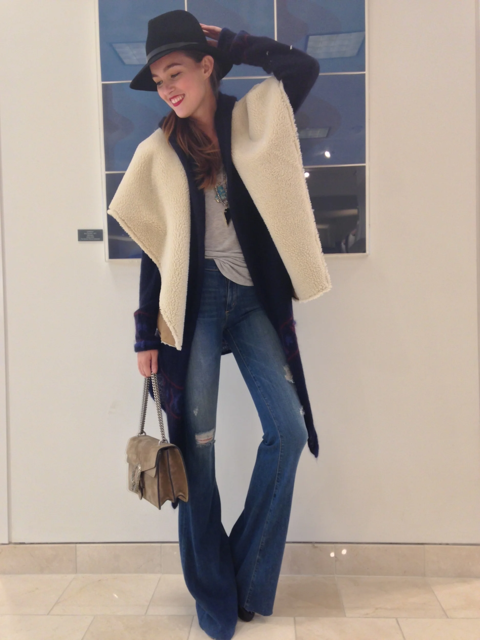 A woman dressed in a casual outfit with a beige fleece vest, blue jeans, and a black hat, posing indoors against a wall with large glass windows.