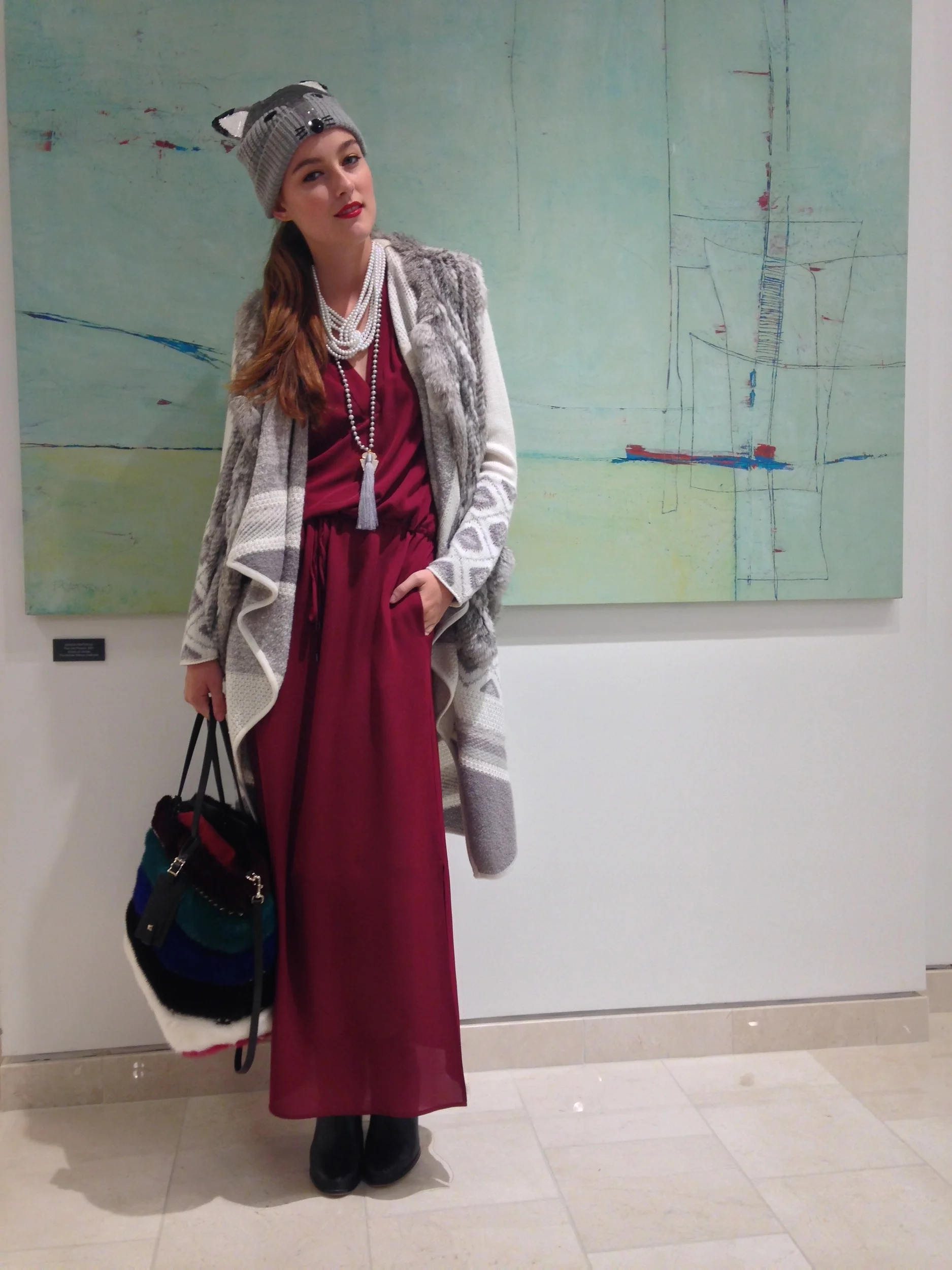 A woman standing in front of an abstract green painting, wearing a animal-themed gray knit hat, multiple pearl necklaces, a red dress, and a long coat with gray and white patterns, holding a black bag with colorful sections.