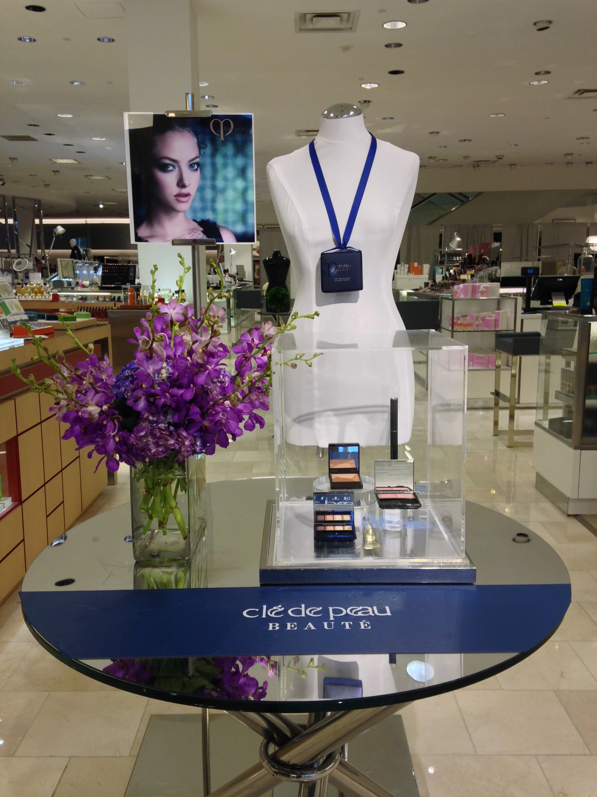 Display table with purple orchids, makeup products, a mannequin with a blue lanyard and a promotional sign for Clé de Peau Beauté in a retail store.