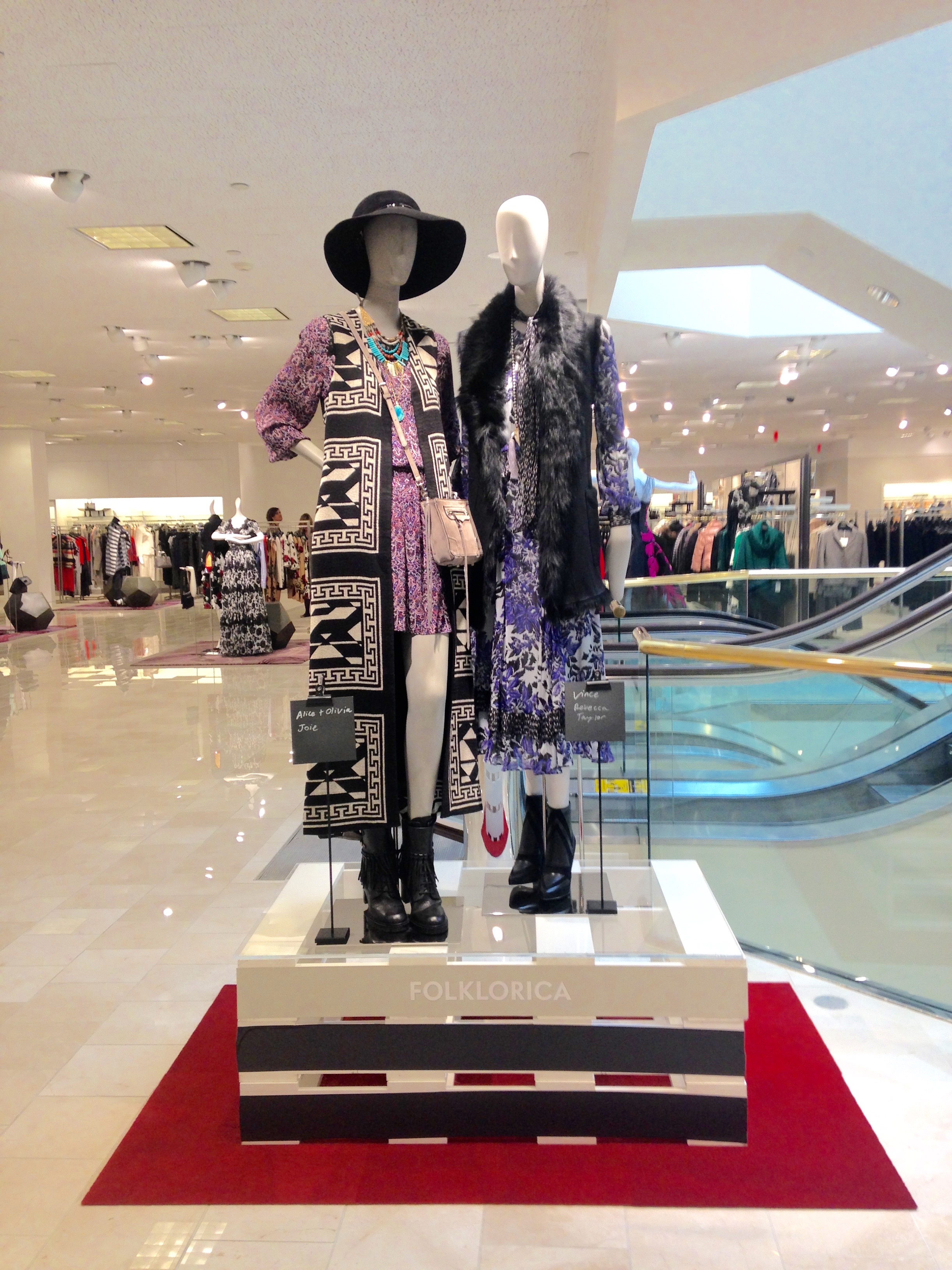 Display of mannequins dressed in bohemian style fashion with one mannequin wearing a hat and layered jewelry, situated on a platform with a red carpet under it, inside a retail store.