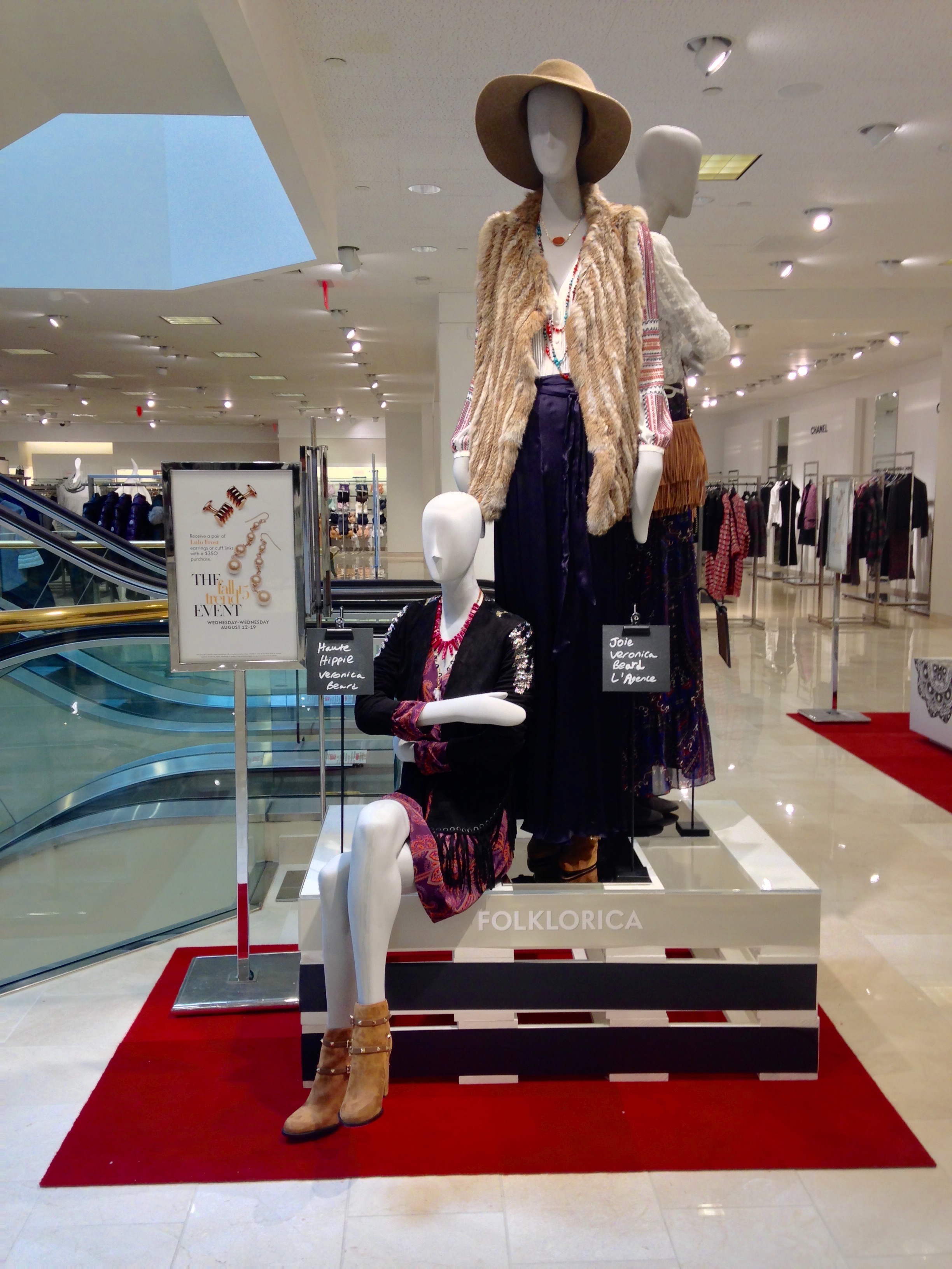 Display of mannequins wearing vintage-inspired women's clothing, including a wide-brimmed hat, fur vest, patterned blouses, long skirts, and high-heeled boots, in a retail store setting with a red carpet and shopping racks in the background.