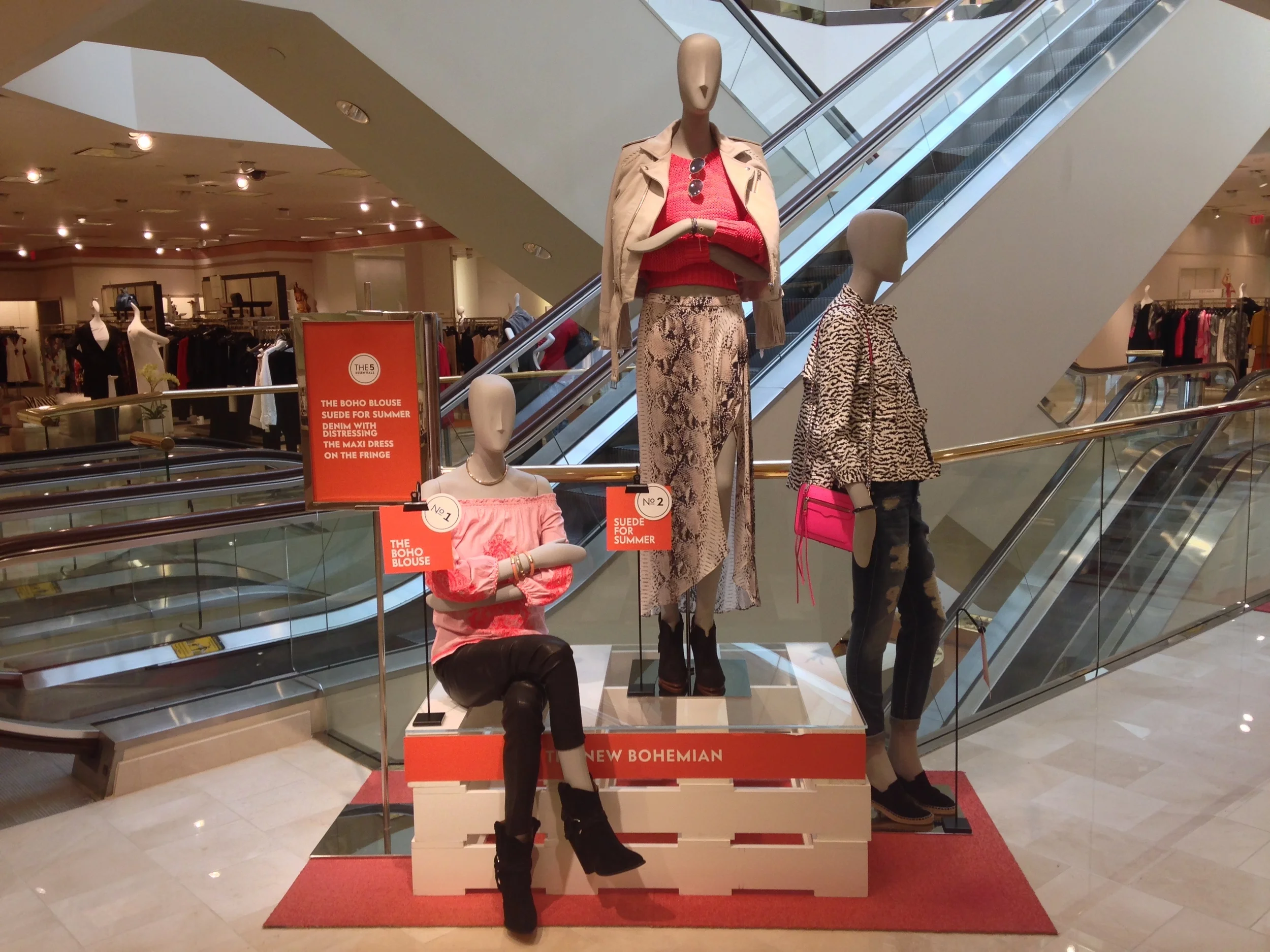 Display of mannequins dressed in casual summer clothing in a clothing store, with escalators and clothing racks in the background.