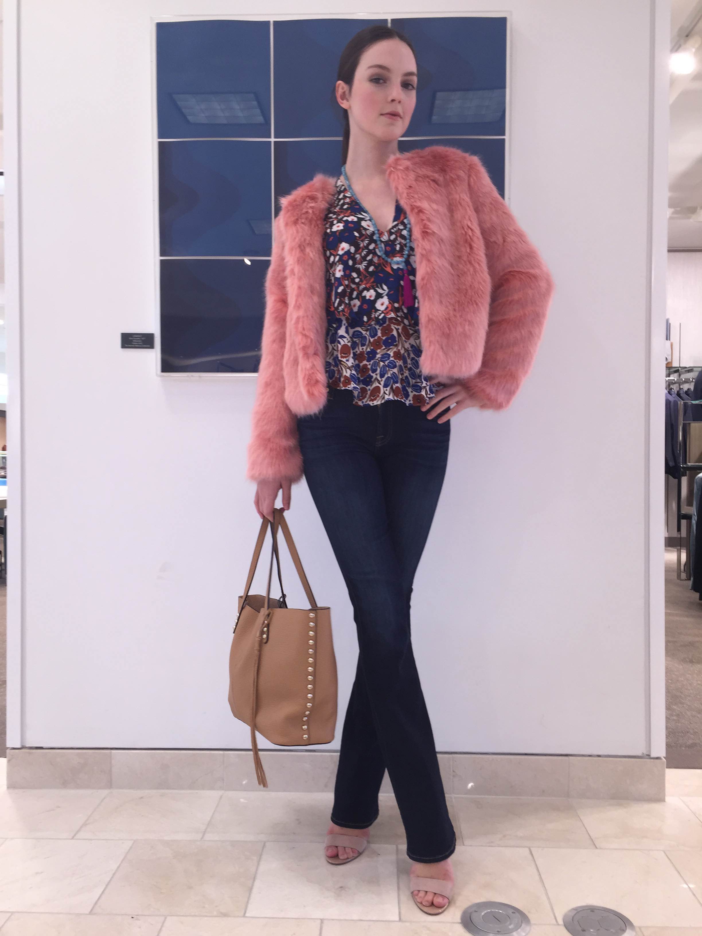 A young woman standing in front of a wall with blue panels, wearing a pink faux fur jacket, a floral blouse, dark jeans, and beige high heels. She is holding a tan handbag with gold studs and has dark hair pulled back. The setting appears to be indoo