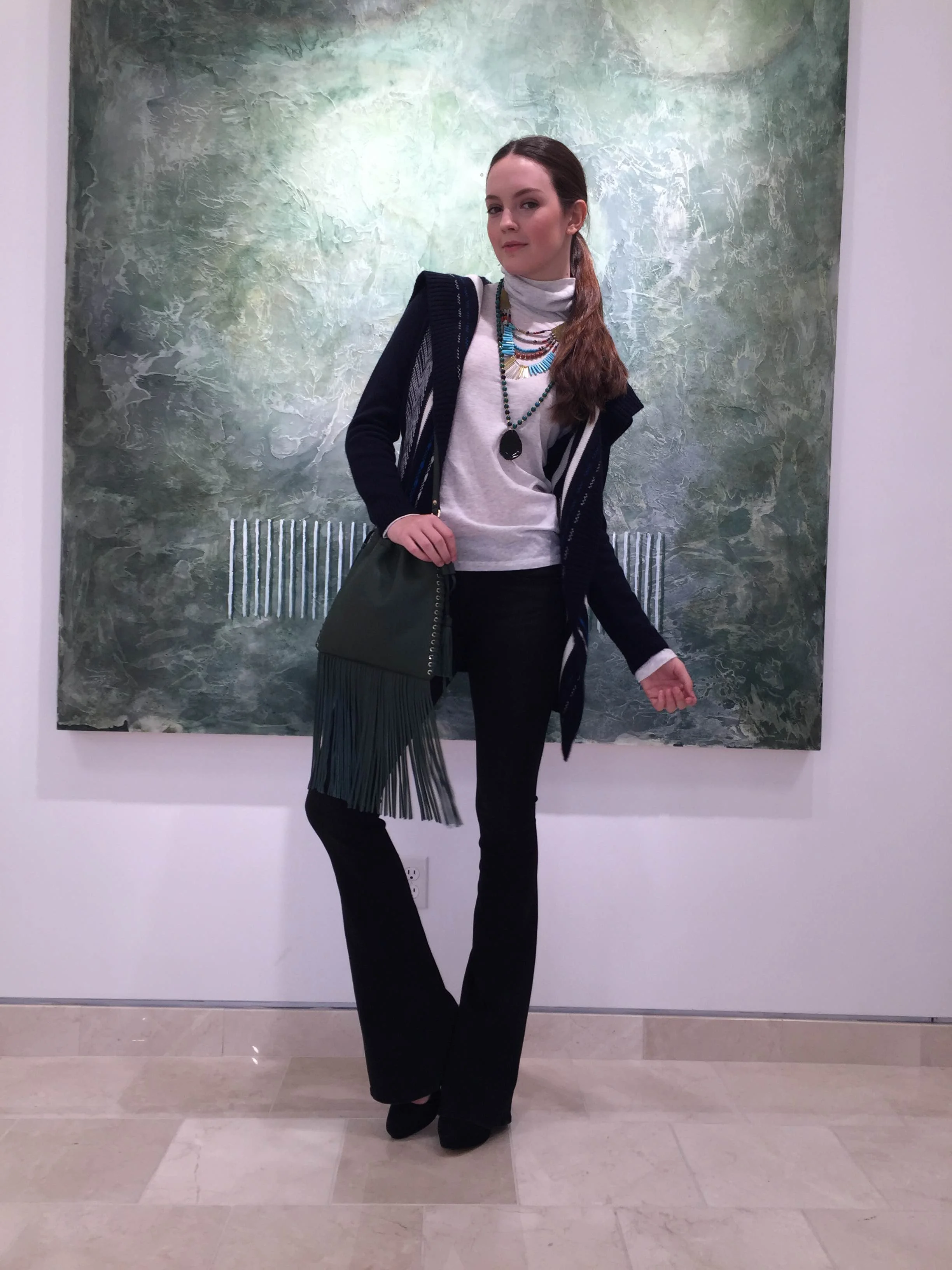 A woman with brown hair in a ponytail stands in front of an abstract green and white textured painting, wearing a white turtleneck, layered necklaces, a black and white cardigan, black pants, and black shoes.
