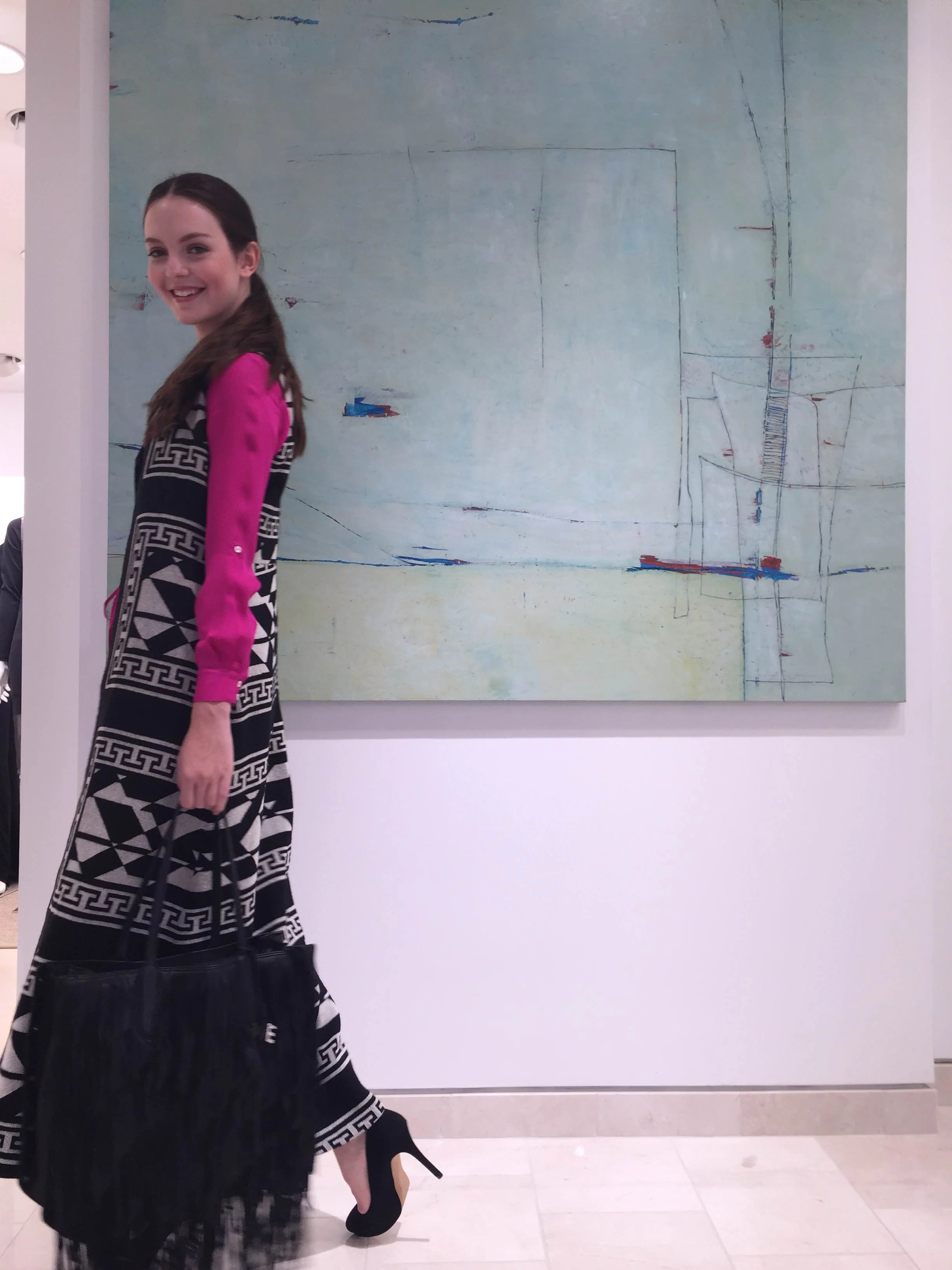 A smiling young woman with brown hair, dressed in a black and white patterned long skirt and a pink long-sleeve top with black designs, stands in front of an abstract painting at an indoor art gallery.