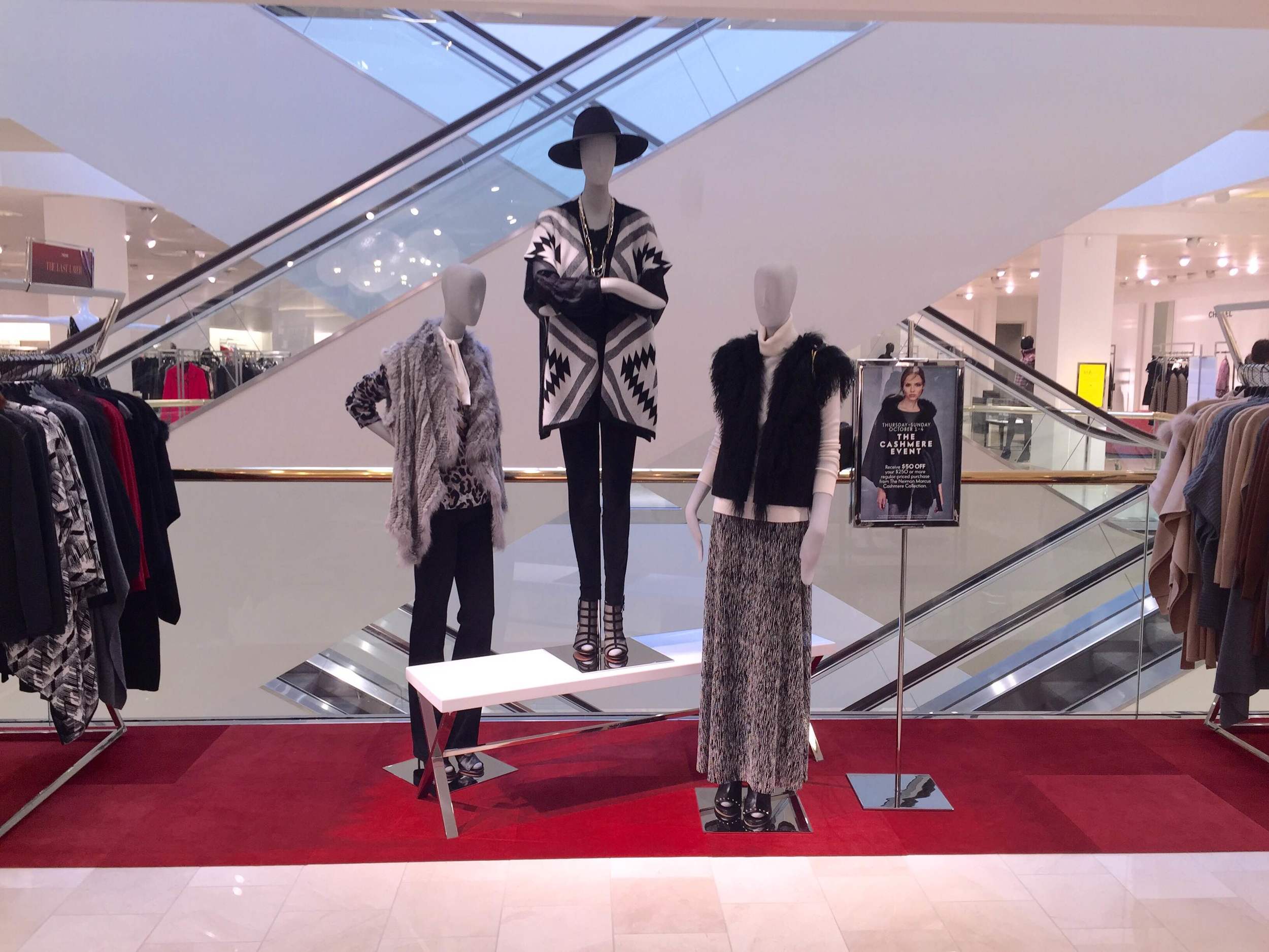 Display of mannequins dressed in fashionable clothing inside a shopping mall store. Two mannequins are dressed in fur and animal print outfits, with one wearing a wide-brimmed hat and jewelry. The mannequins are positioned on a red carpeted platform 