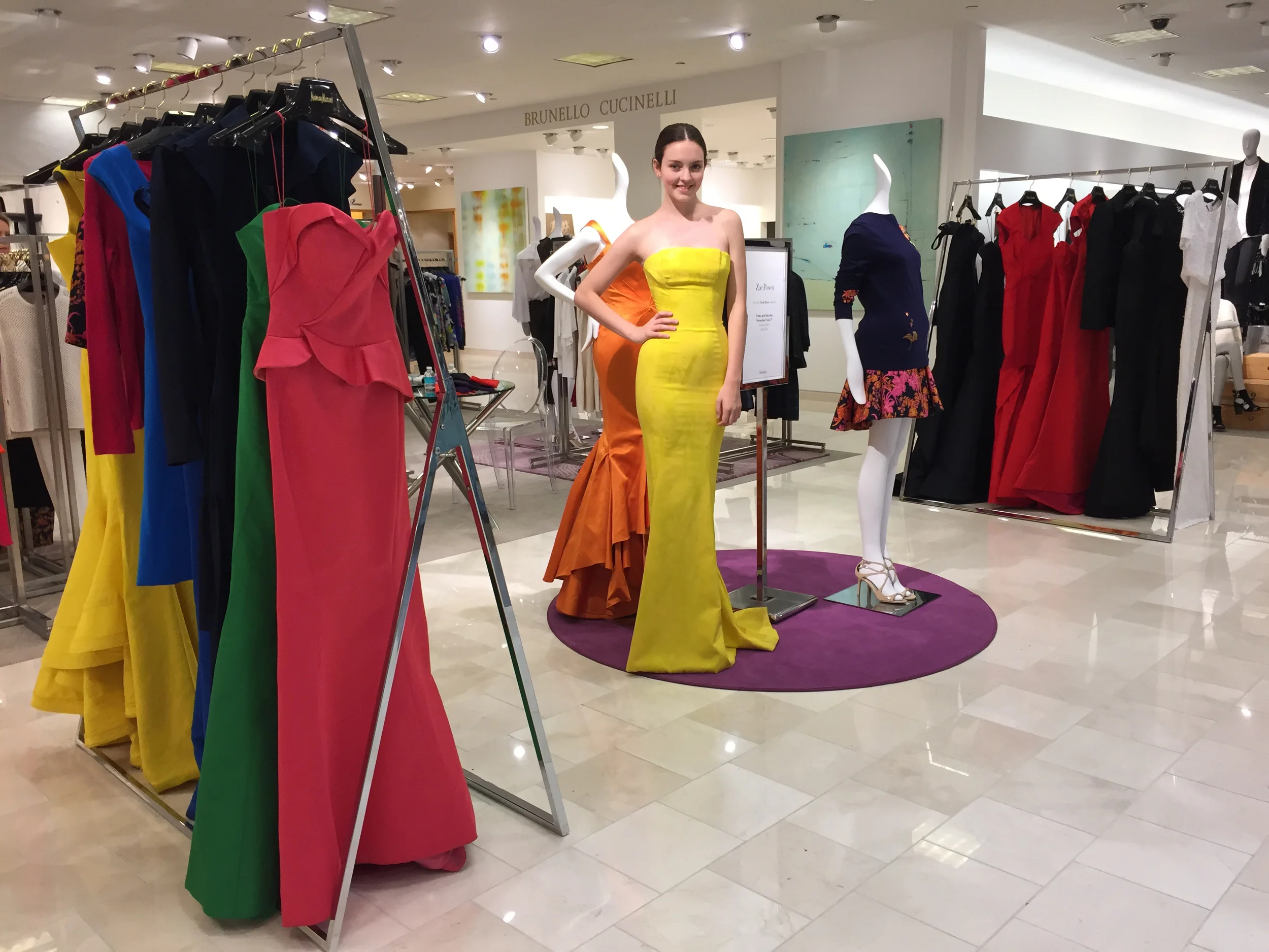 A woman in a strapless yellow dress stands in a clothing store surrounded by racks of colorful dresses and mannequins, with a sign above that reads "Brunello Cucinelli."