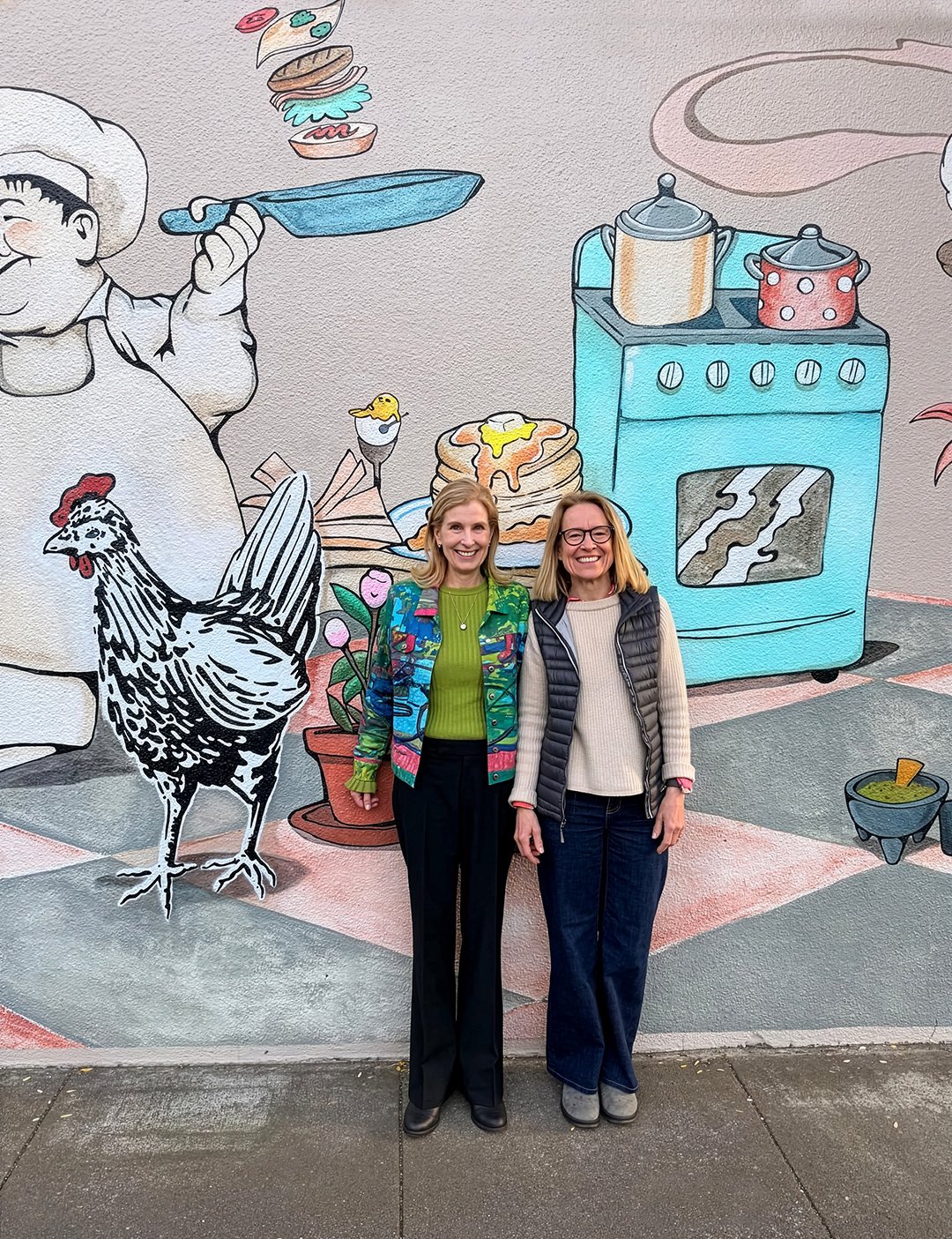 Natasha and Emily Luchetti outside Omnivore beside the vibrant mural featuring characters from "In the Night Kitchen" by artist Josué Rojas.