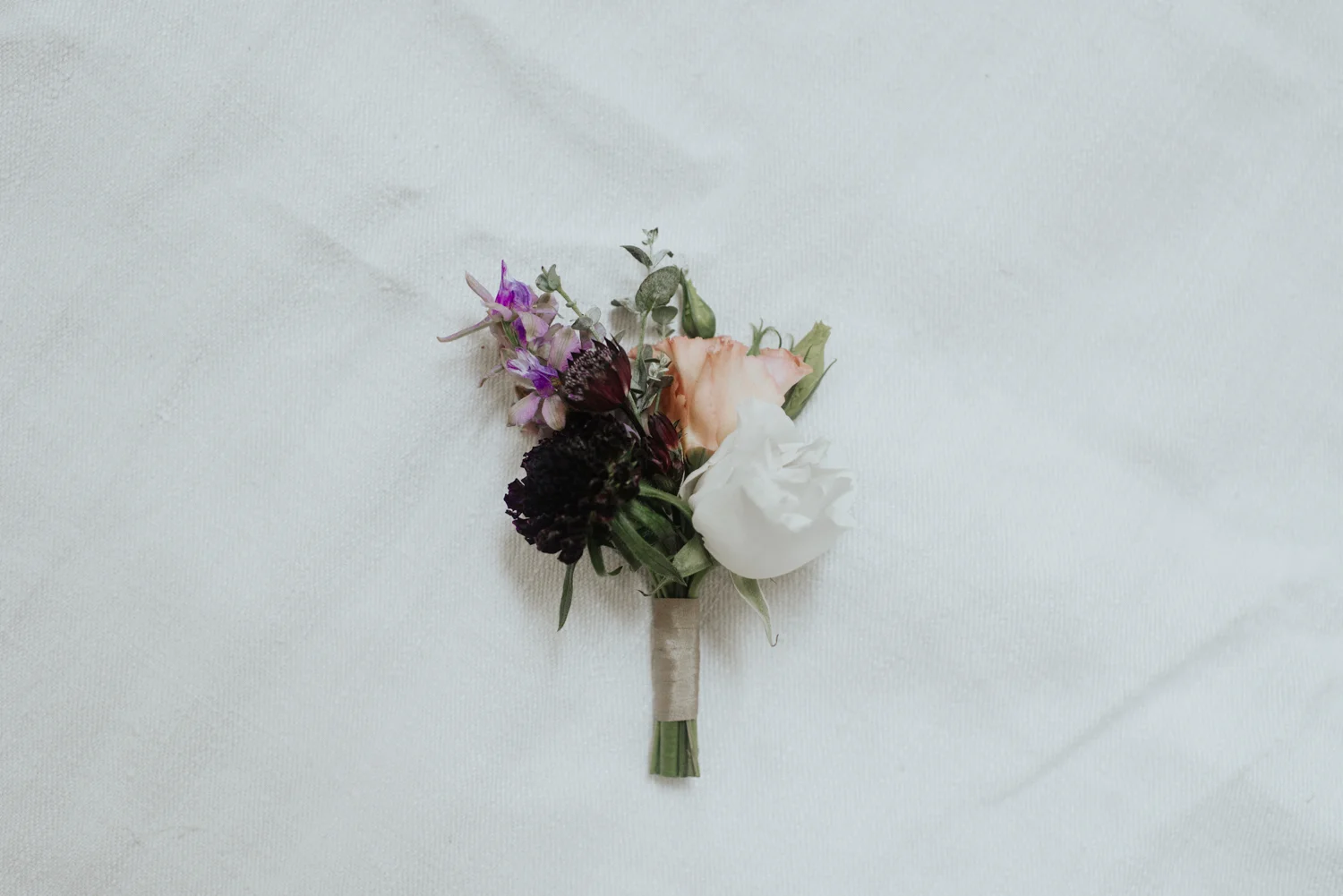 Boutonniere $15 Boutonnieres are well suited for men or women. They are typically worn on jacket lapels, vests or on double layer dresses but we do not recommend these for single layer dresses or shirts. Our boutonnieres are composed of smaller accent flowers as well as greenery and are wrapped with ribbon. Boutonnieres include two pins to affix them. Traditionally grooms, groomsmen, ring bearers, fathers, grandfathers, and ushers wear boutonnieres to signify they are family or a part of the wedding party. Ladies who prefer to wear a flower over having one on their wrist may also wear boutonnieres. For our boutonnieres, clients have the option of choosing between a primary and secondary color palette for the flowers as well as a ribbon color. Color palette and ribbon color choices will remain consistent throughout all bouquets, boutonnieres, and corsages ordered. In this example, our client chose a lavender/mauve primary color palette with a blush secondary color palette and a nude silk ribbon.
