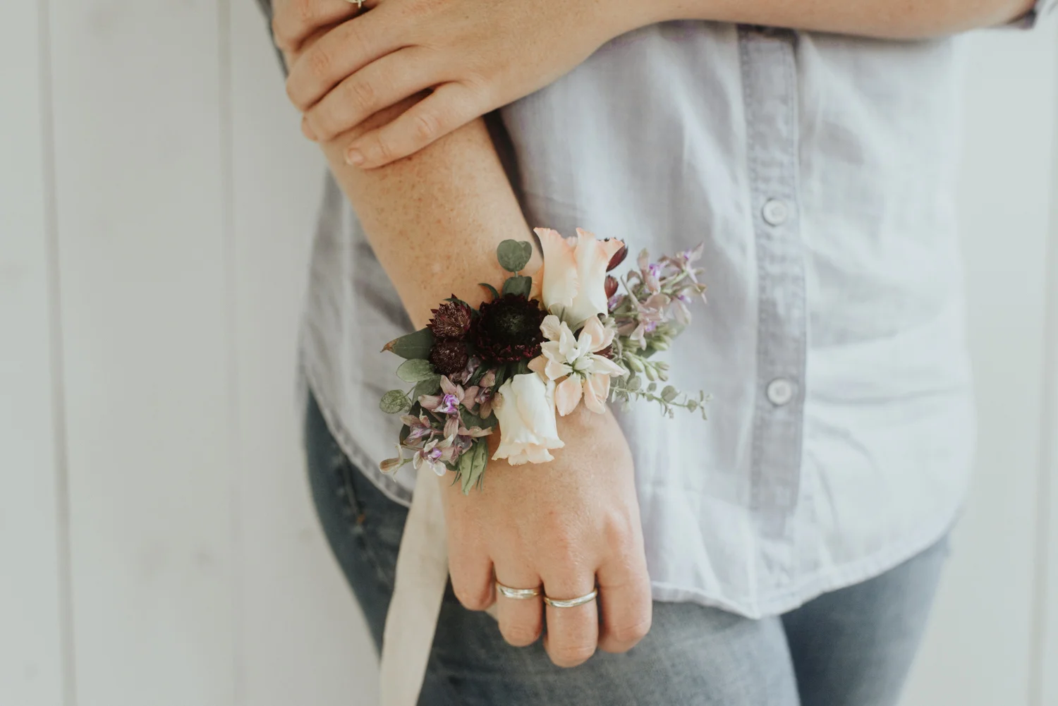 Corsage $30 Corsages are great for mothers, grandmothers, and bridal parties. Our corsages are composed of flowers and greenery and include the choice of a ribbon that ties comfortably around the wrist in the classic style or an adjustable metal cuff bracelet base for a more modern look. For our corsages, clients have the option of choosing between a primary and secondary color palette for the flowers as well as a ribbon color or the cuff base option. Color palette and ribbon color choices will remain consistent throughout all bouquets, boutonnieres, and corsages ordered. In this example, our client chose a lavender/mauve primary color palette with a blush secondary color palette and a blush silk ribbon.