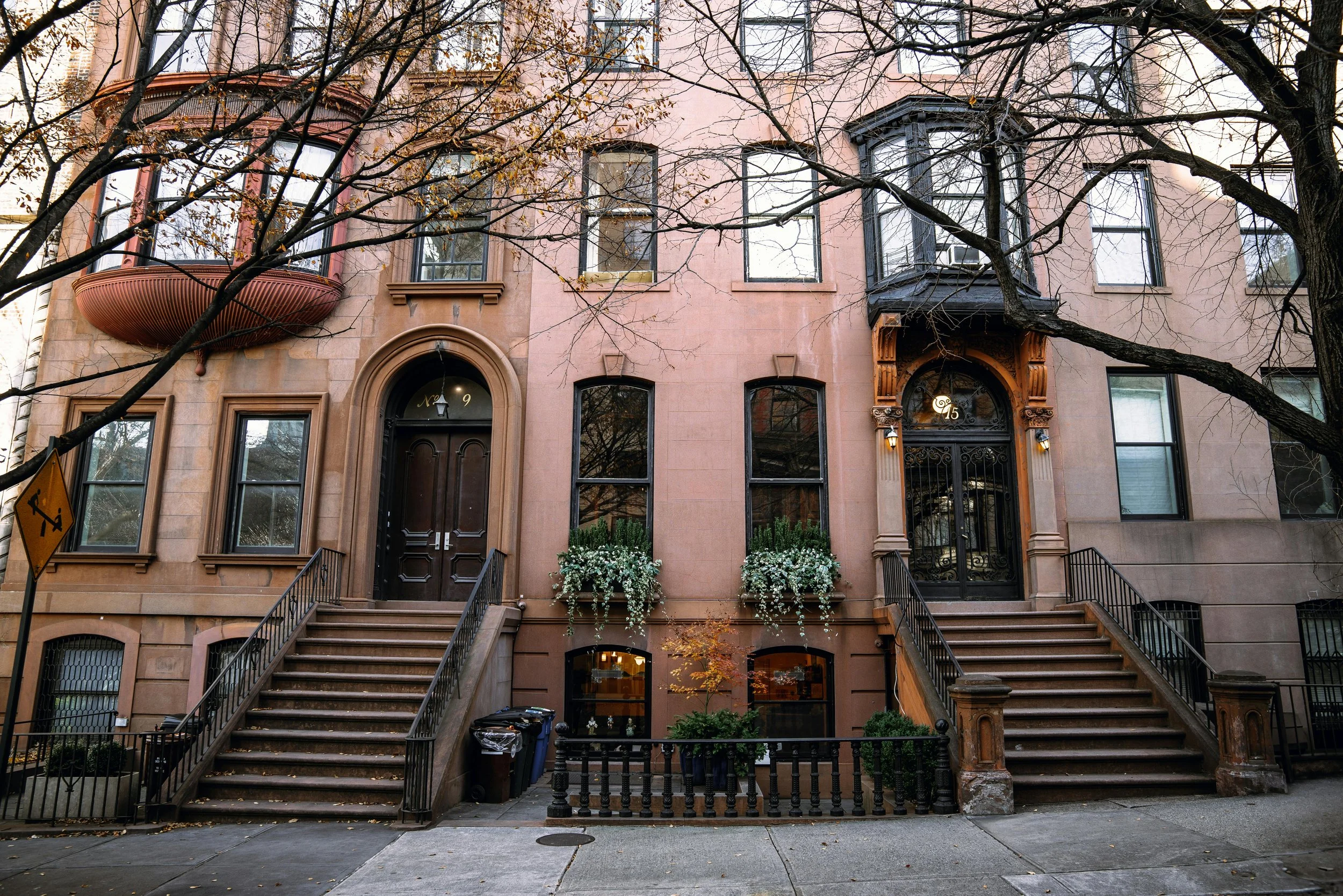 Brownstone row house with stairs leading to two front doors, surrounded by trees with some leaves fallen on sidewalk.