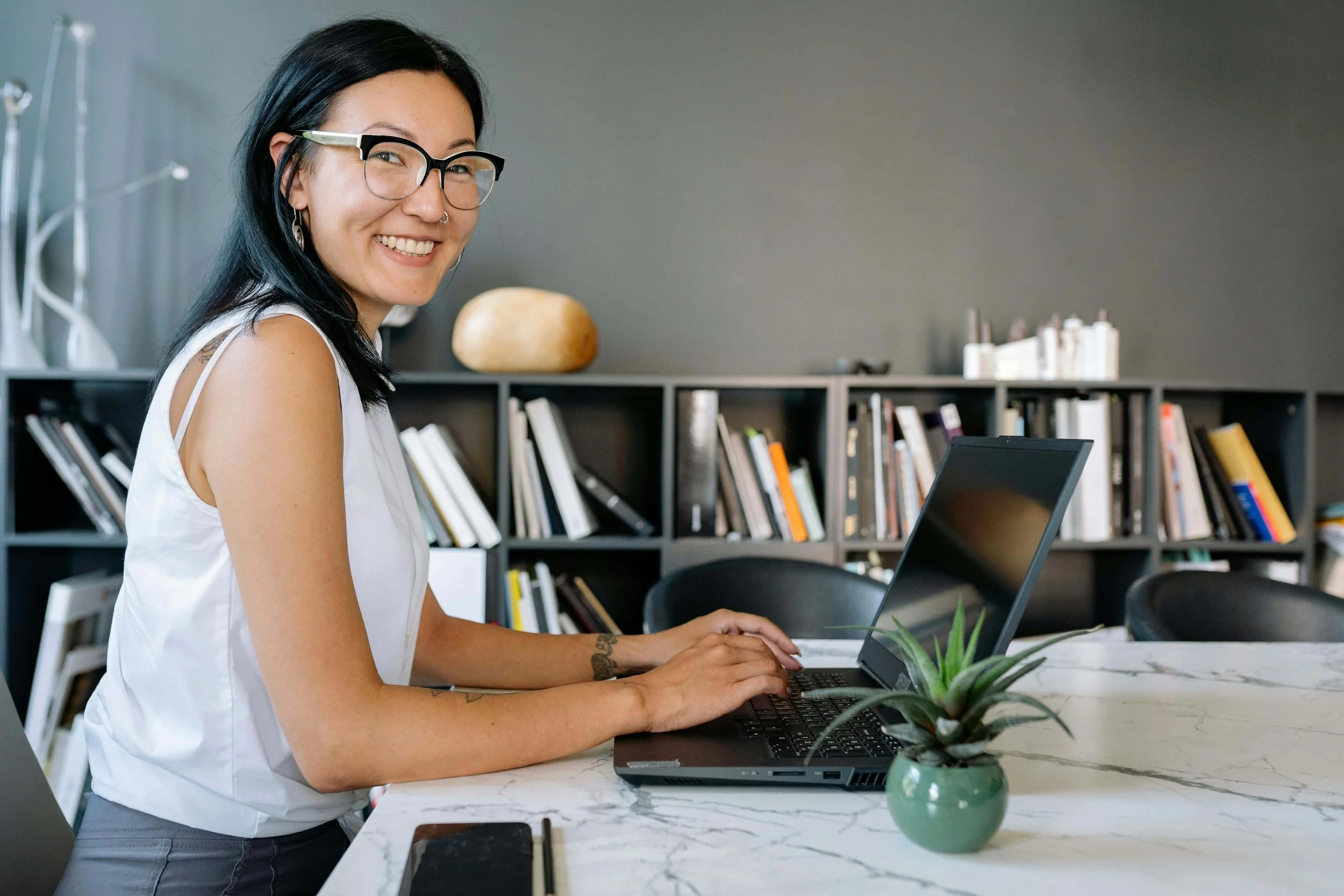 Smiling woman with glasses working on a laptop at a white marble table, with a bookshelf filled with books in the background.