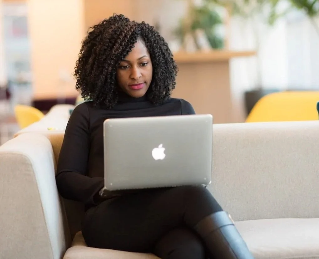 A woman with curly black hair sitting on a beige couch, using a silver Apple MacBook laptop in a well-lit indoor space.