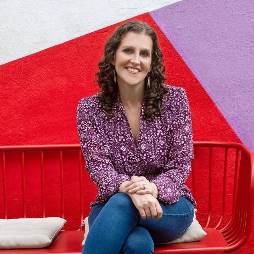A woman with curly brown hair, wearing a purple patterned blouse and blue jeans, sitting on a red bench with white cushions, smiling in front of a colorful wall with red, purple, and white sections.