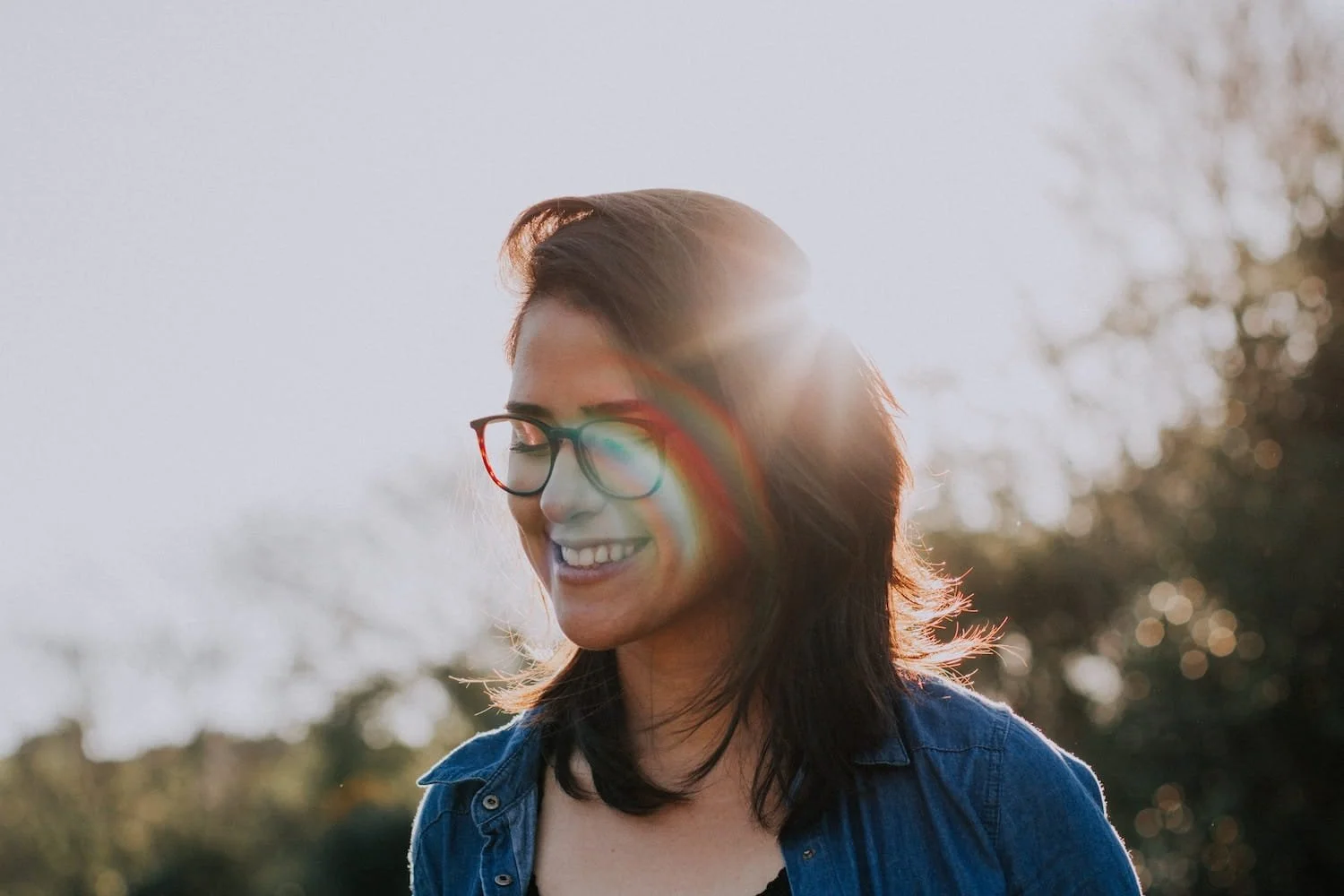 A woman with glasses smiling outdoors in sunlight.