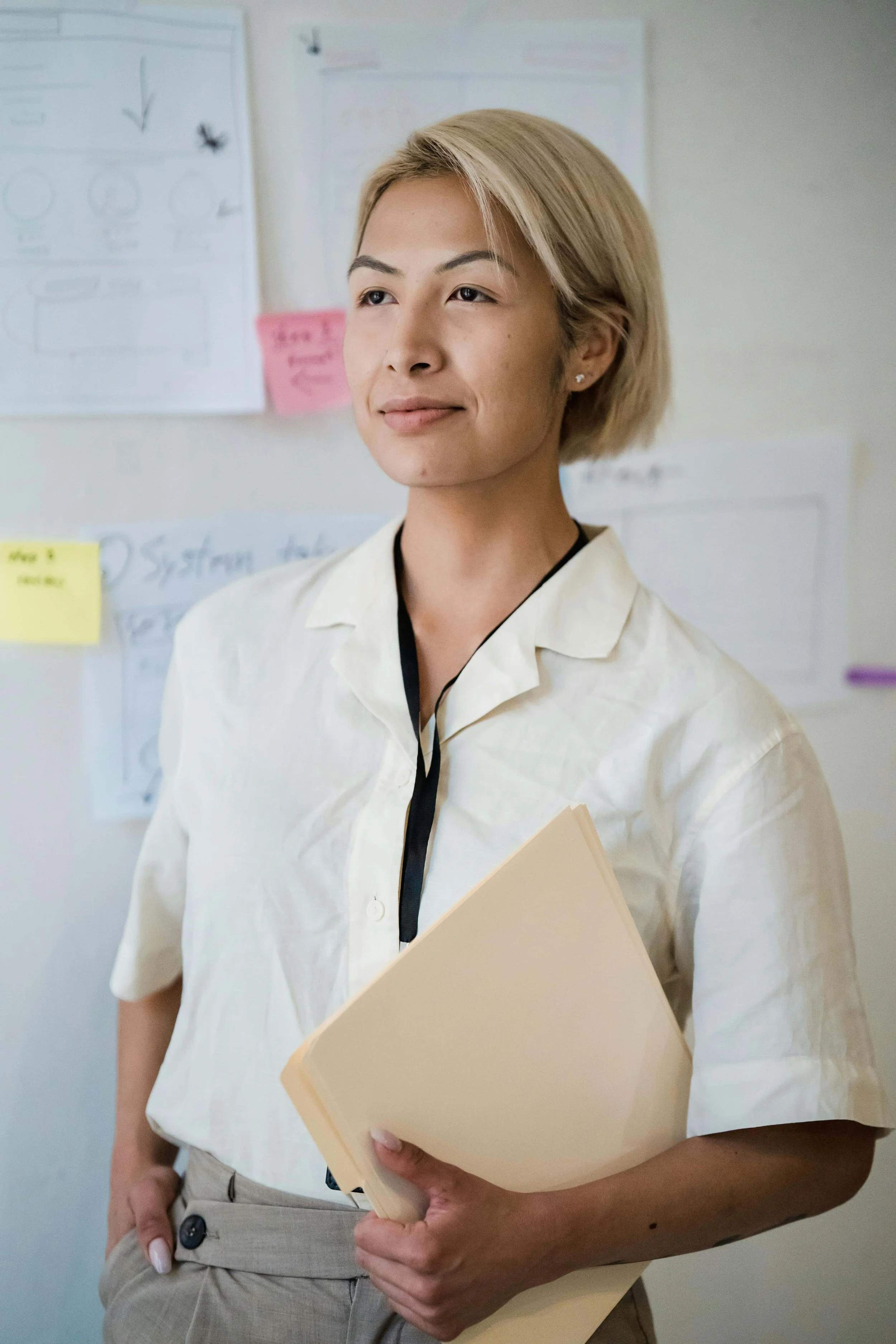 A woman with blonde hair holding a beige folder, standing in front of a whiteboard with notes, charts, and sticky notes.