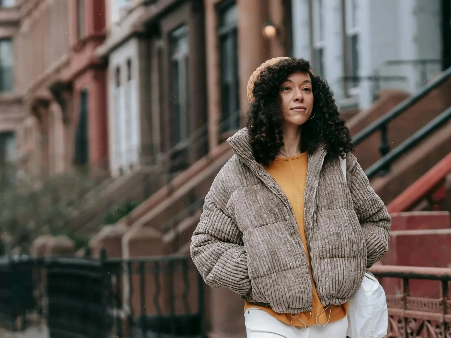 A young woman with curly hair wearing a beige knit hat, brown puffer jacket, yellow shirt, and white pants walking on a city street.