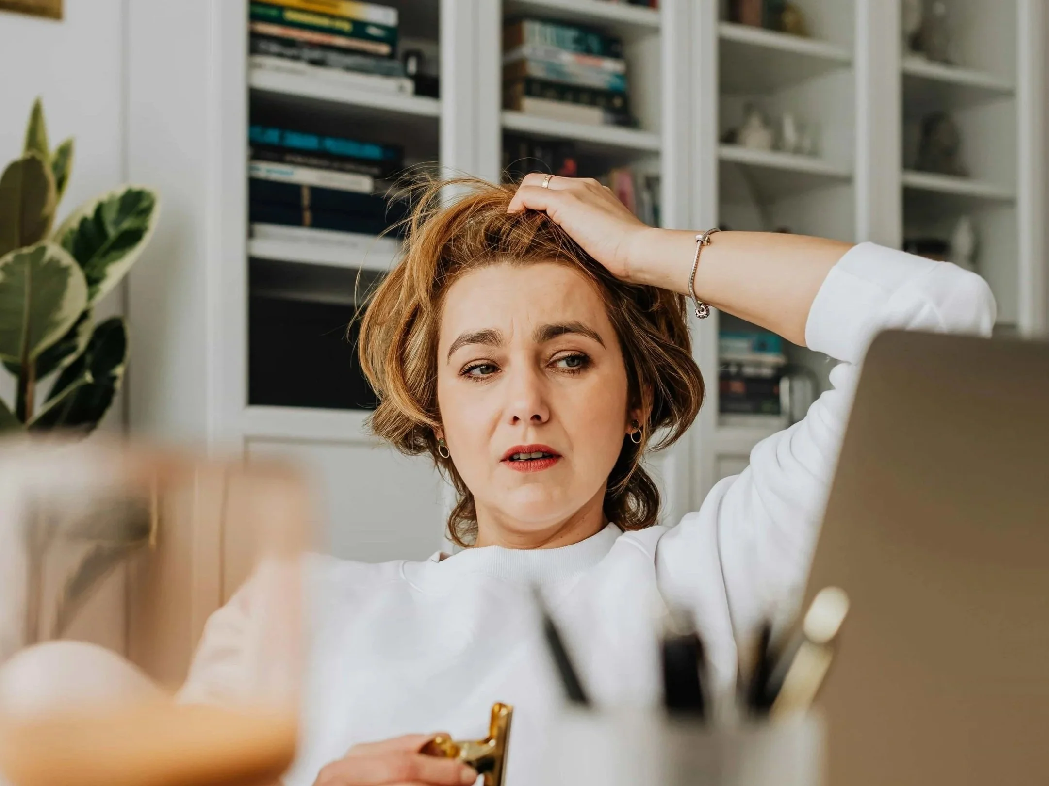 A woman with short, wavy, brown hair and a white shirt sits at a desk, looking stressed or worried, with her hand on her head, in a room with bookshelves and a potted plant.