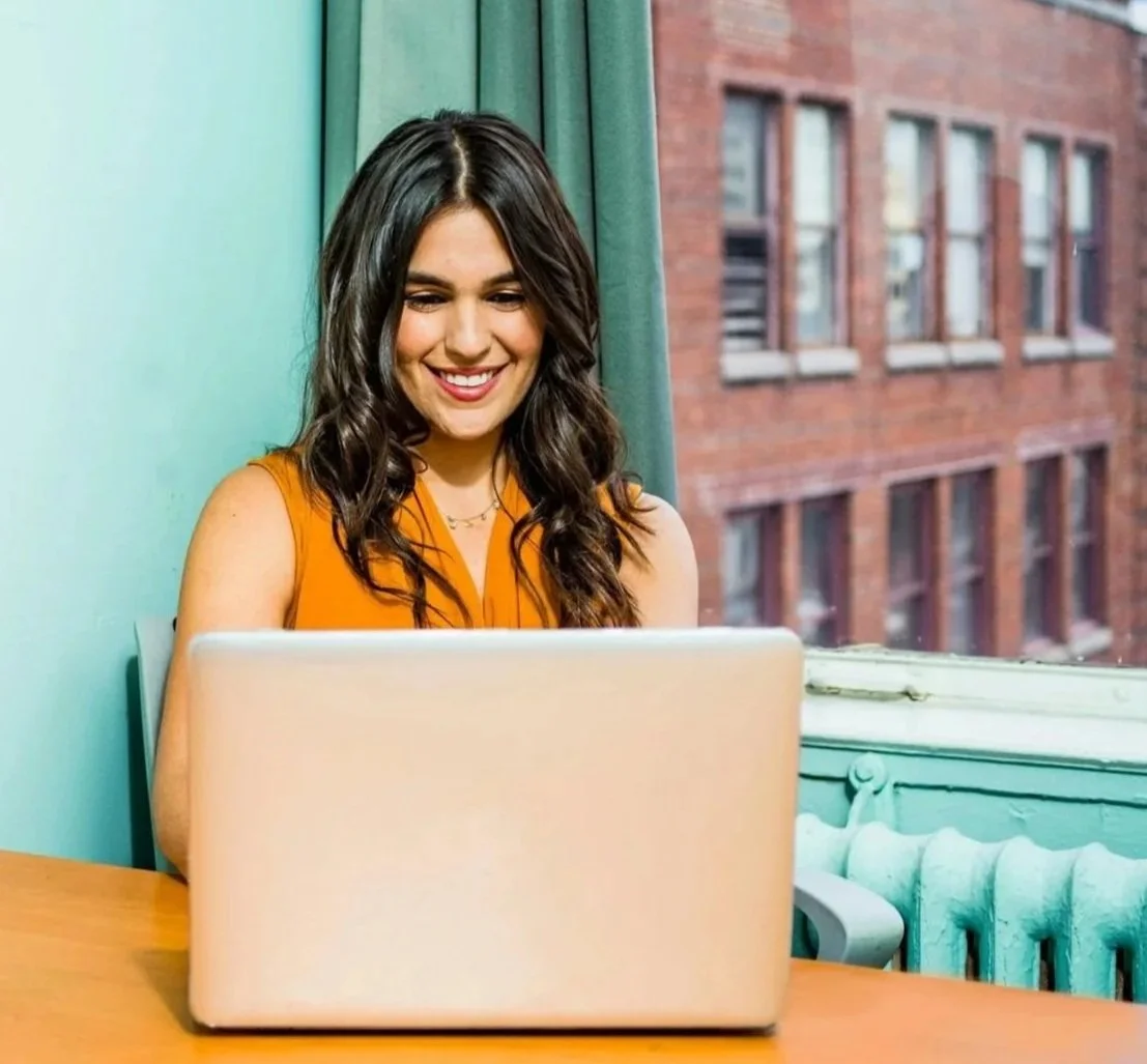 A woman with long dark hair smiling while looking at a laptop in a brightly lit room with a large window showing a brick building outside.