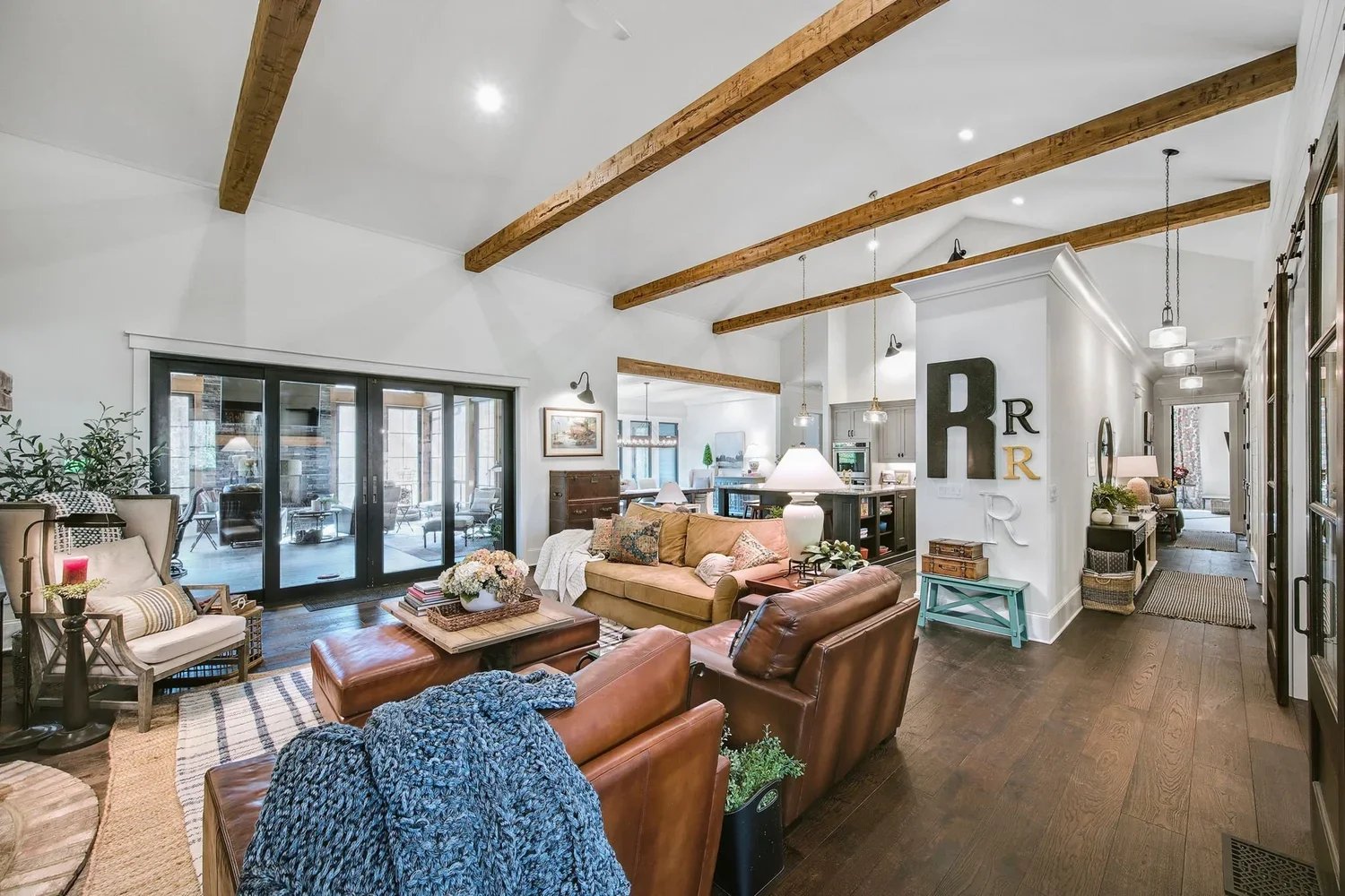 Spacious living room with exposed wooden beams, large sliding glass doors, a mix of leather and fabric sofas, wooden coffee table, decorative pillows, wall art, and a view into a bright kitchen and dining area.