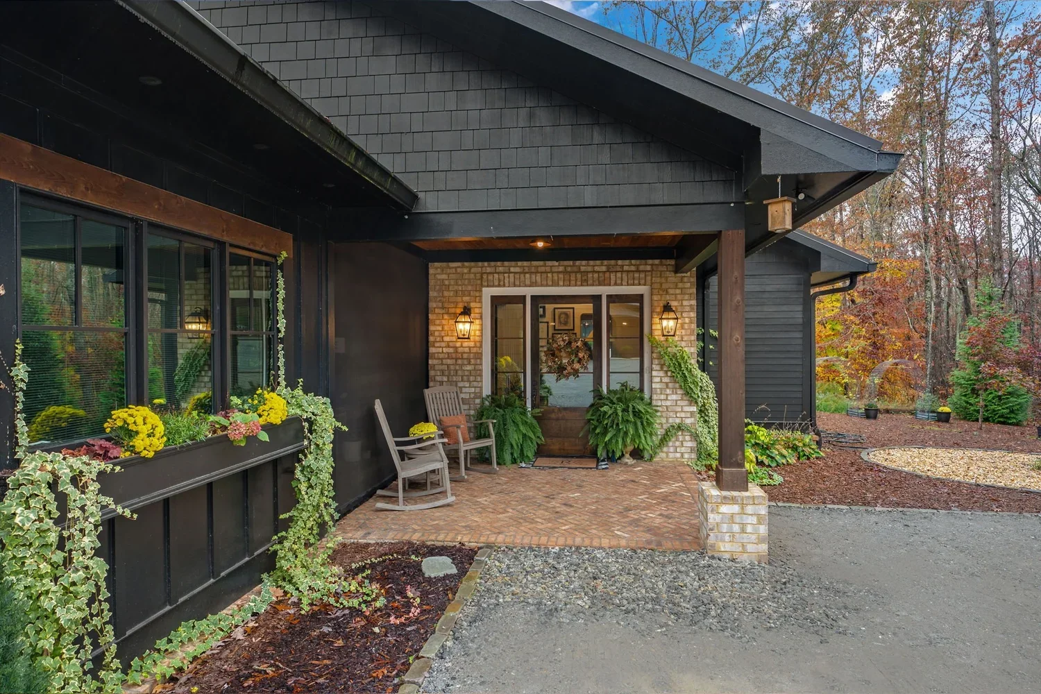 Covered front porch of a house with brick and black siding, decorated with potted plants, hanging lanterns, and two wooden chairs, surrounded by trees with autumn foliage.