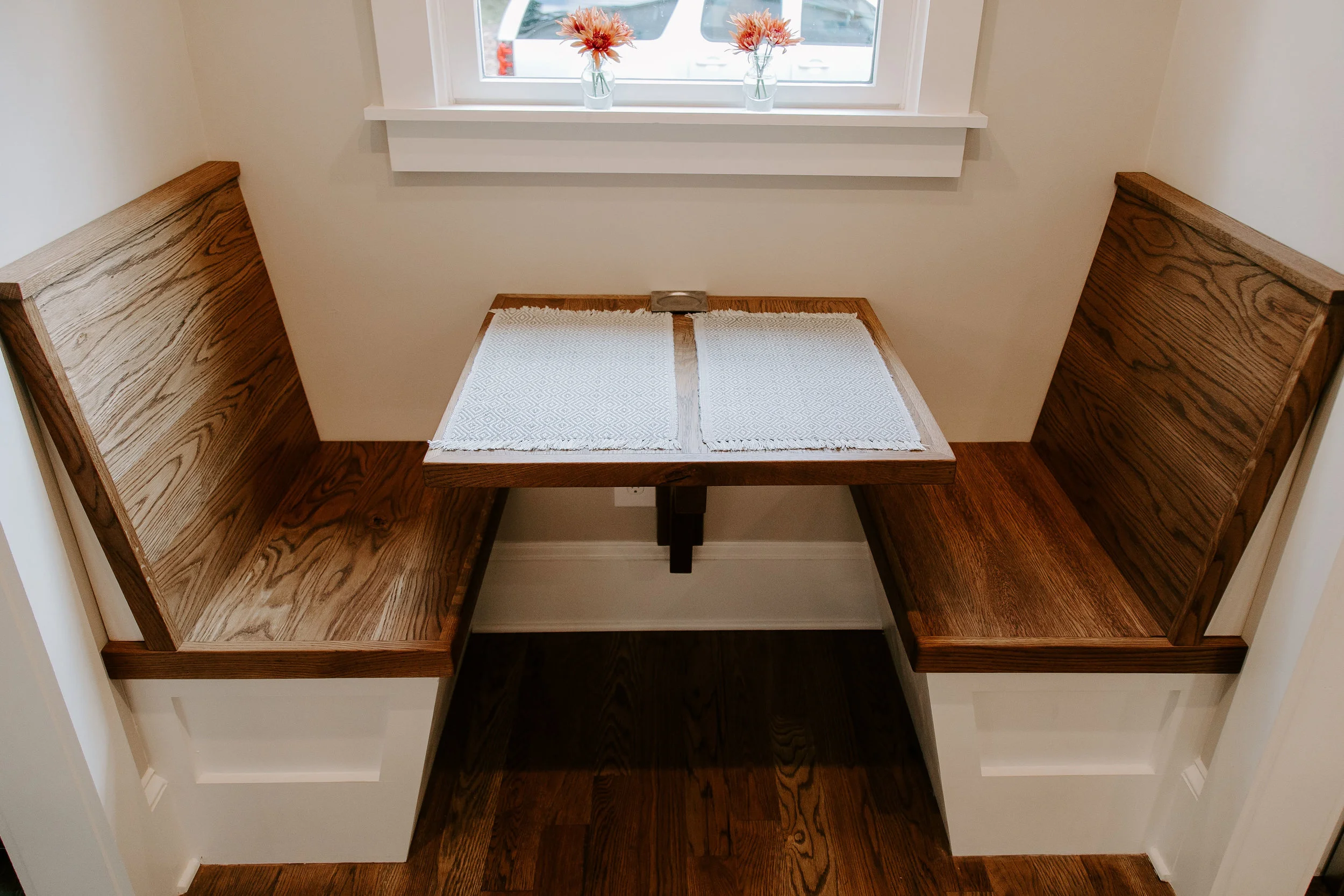 Breakfast nook with built-in wood bench seating, a wall-mounted solid wood dining table, rich dark hardwood flooring, and a white-trimmed window above.
