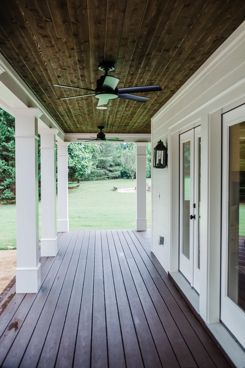 Covered outdoor porch with a dark stained wood plank ceiling, two black ceiling fans, white square support columns, brown composite deck flooring, and white French doors with glass panels.
