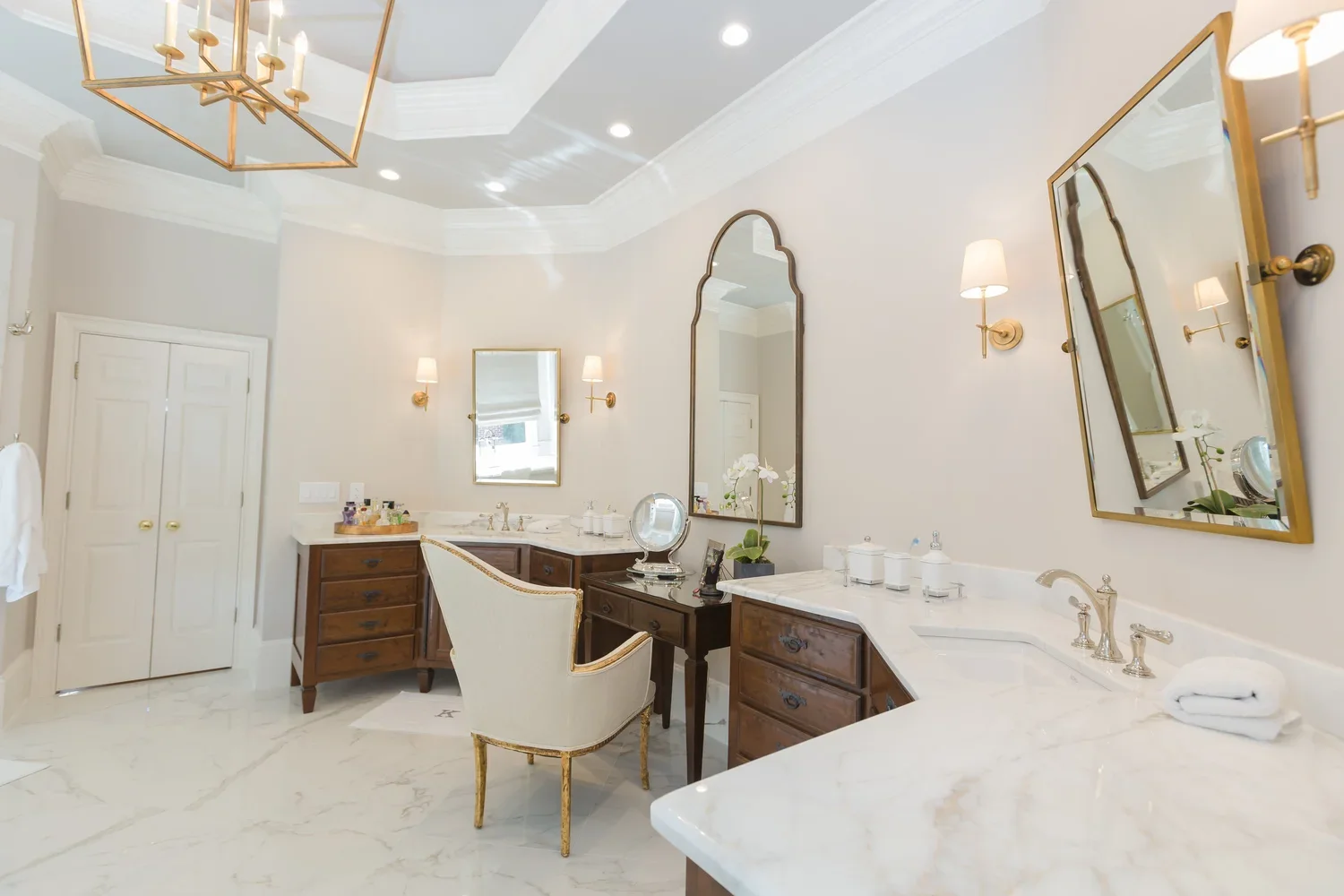 Elegant bathroom with marble countertops, wooden cabinets, large mirrors framed in gold, and wall-mounted lamps with white shades