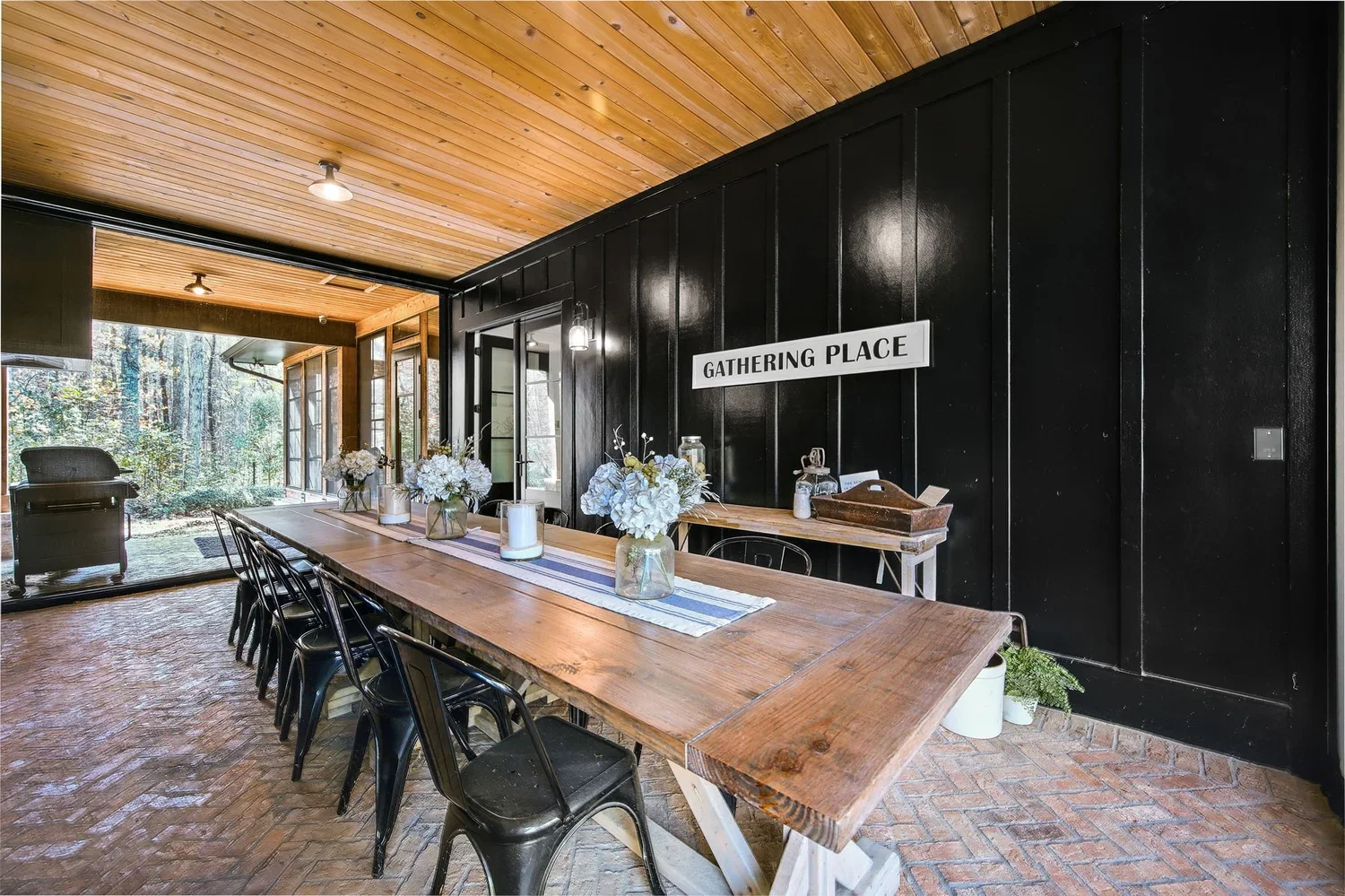 Indoor gathering space with a long wooden table decorated with flower arrangements and candles, black chairs, black paneled wall with sign 'Gathering Place', and a view of a screened porch with outdoor furniture and trees outside.