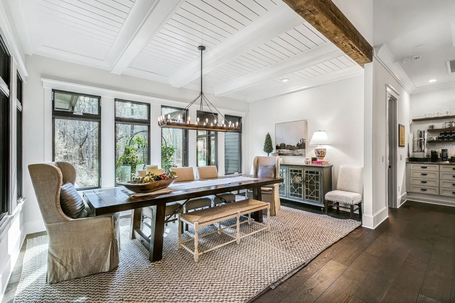 Dining room with wooden table, six chairs, beige upholstered armchair, white and beige bench, chandelier, large windows, and greenery outside. Decor includes a sideboard with a lamp, artwork, and a rug.