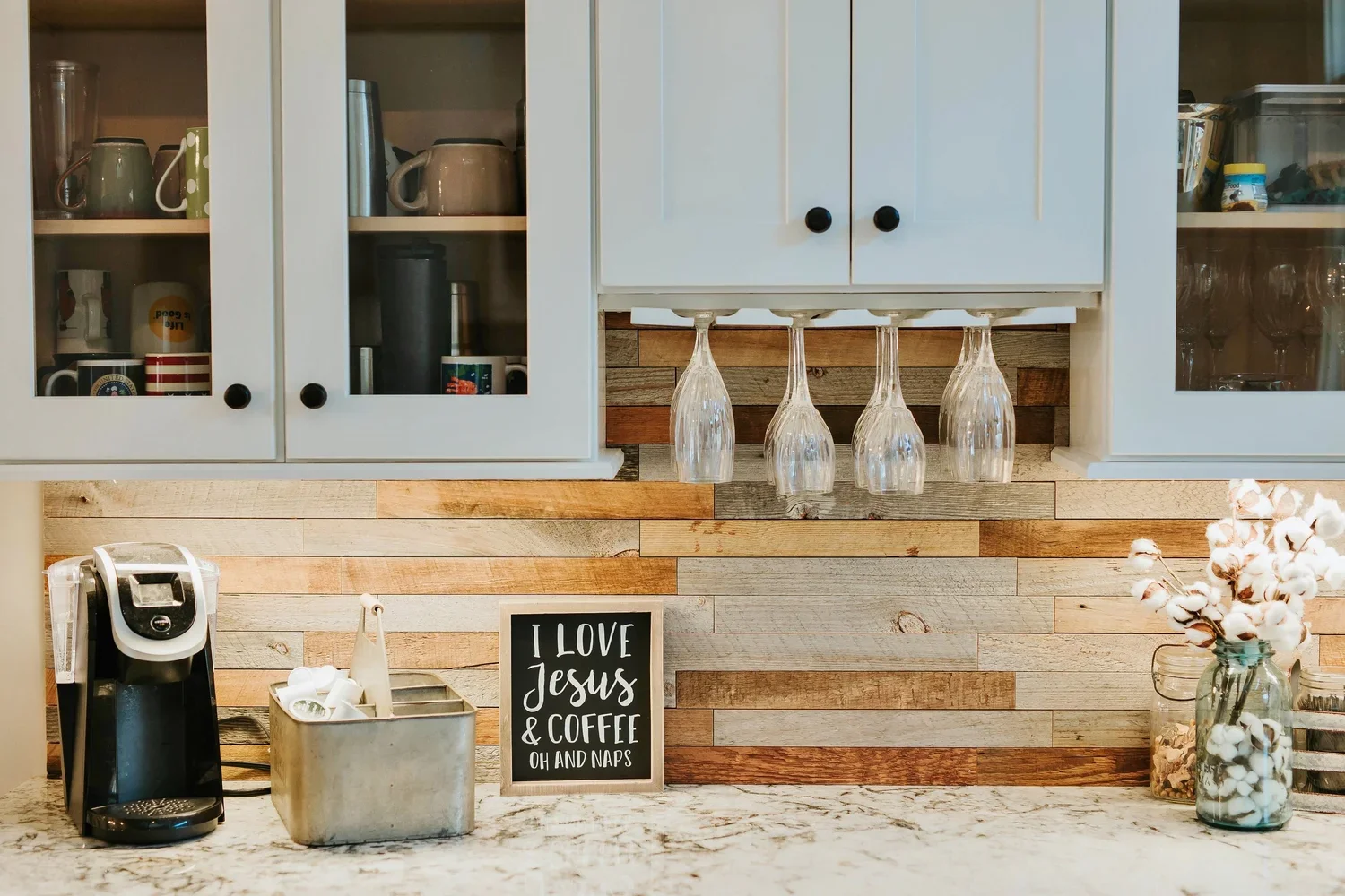 Kitchen countertop with coffee machine, metal container with coffee pods, a small sign that says "I love Jesus & coffee, oh and naps," glassware hanging upside down, and a vase with cotton flowers.