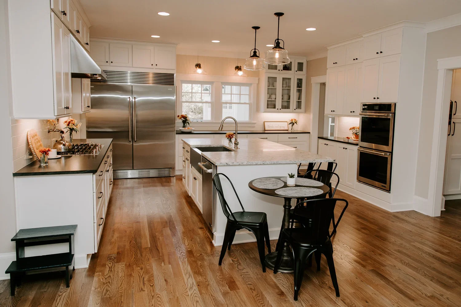 Kitchen with ceiling-height white shaker cabinetry, a large central island with a light stone countertop and built-in sink, stainless steel appliances, white tile backsplash, built-in wall ovens, and wide-plank medium-tone hardwood flooring.