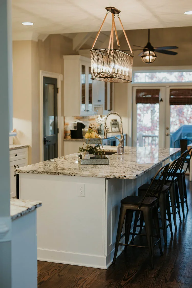 Modern kitchen with an oversized waterfall-style white-and-gray veined quartz island, integrated undermount sink, dark hardwood flooring, and oversized industrial pendant lighting, extended island seating, white shaker-style cabinetry, and large glas