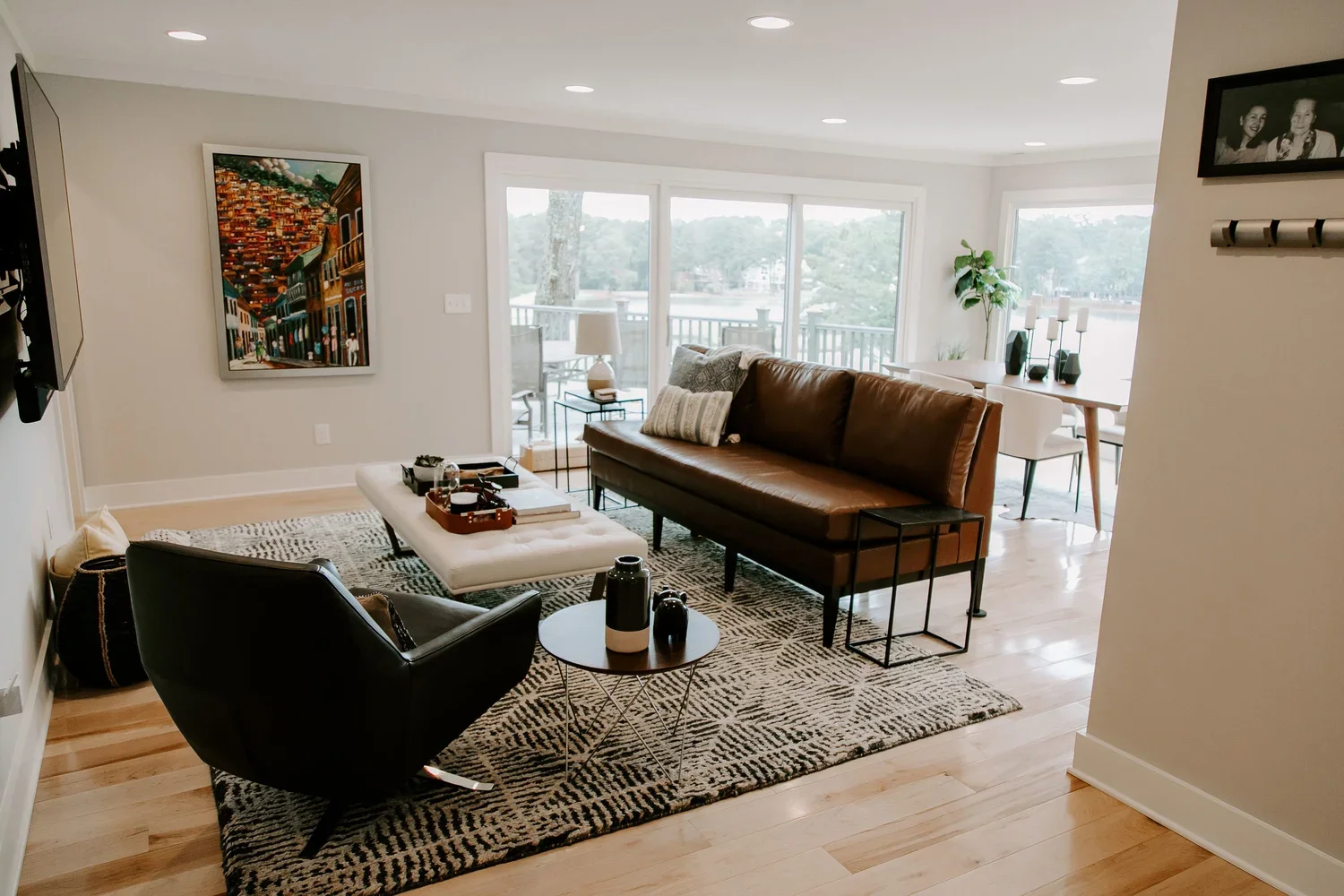 Living room with a brown leather sofa, a black armchair, a white ottoman, a patterned rug, art on the wall, and large windows overlooking a deck and trees.