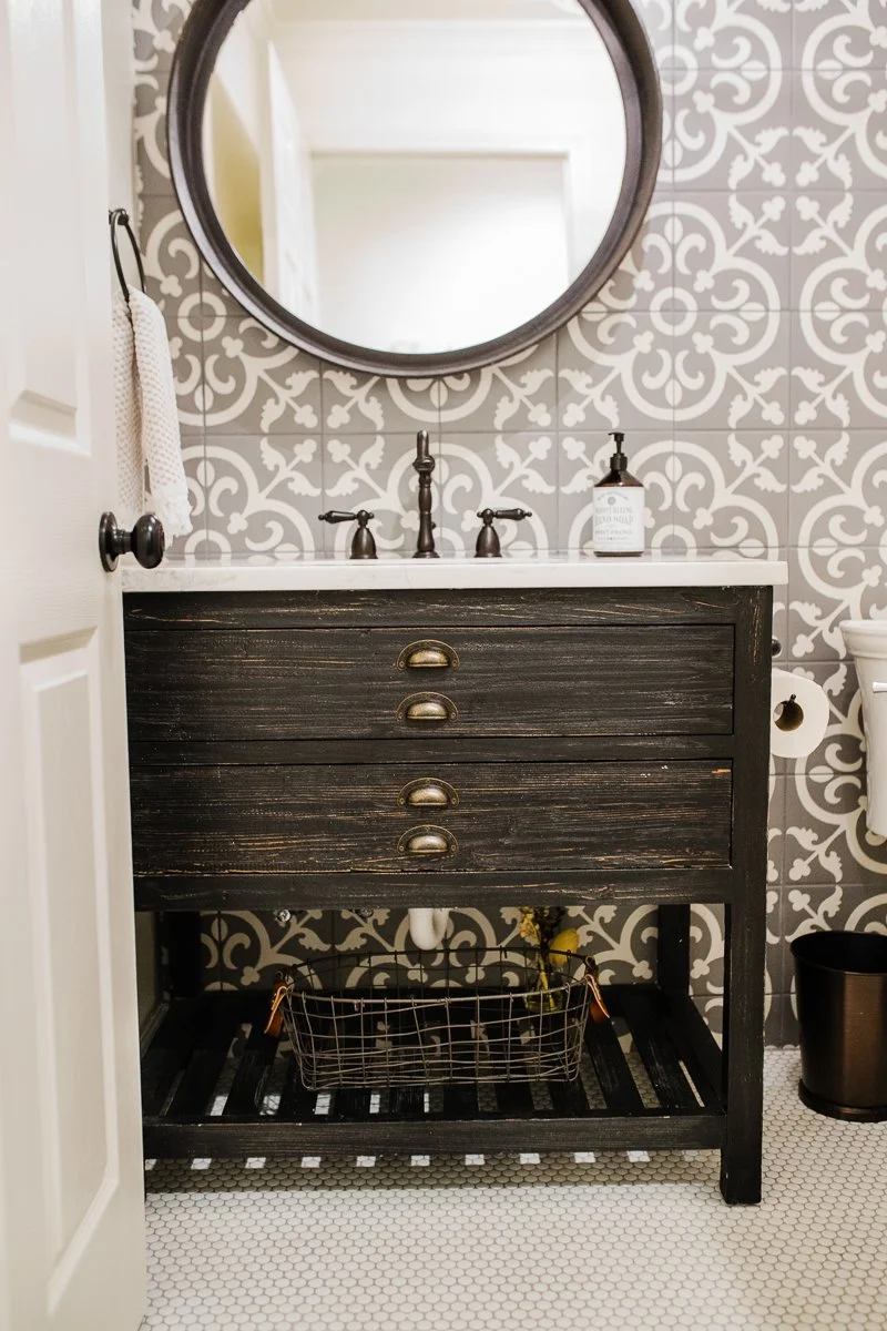 Bathroom vanity with dark wood finish, three drawers with vintage handles, a white countertop, and a round mirror above. The wall has gray and white patterned tiles. There is a soap dispenser on the countertop, a towel hanging on the wall, and a wire