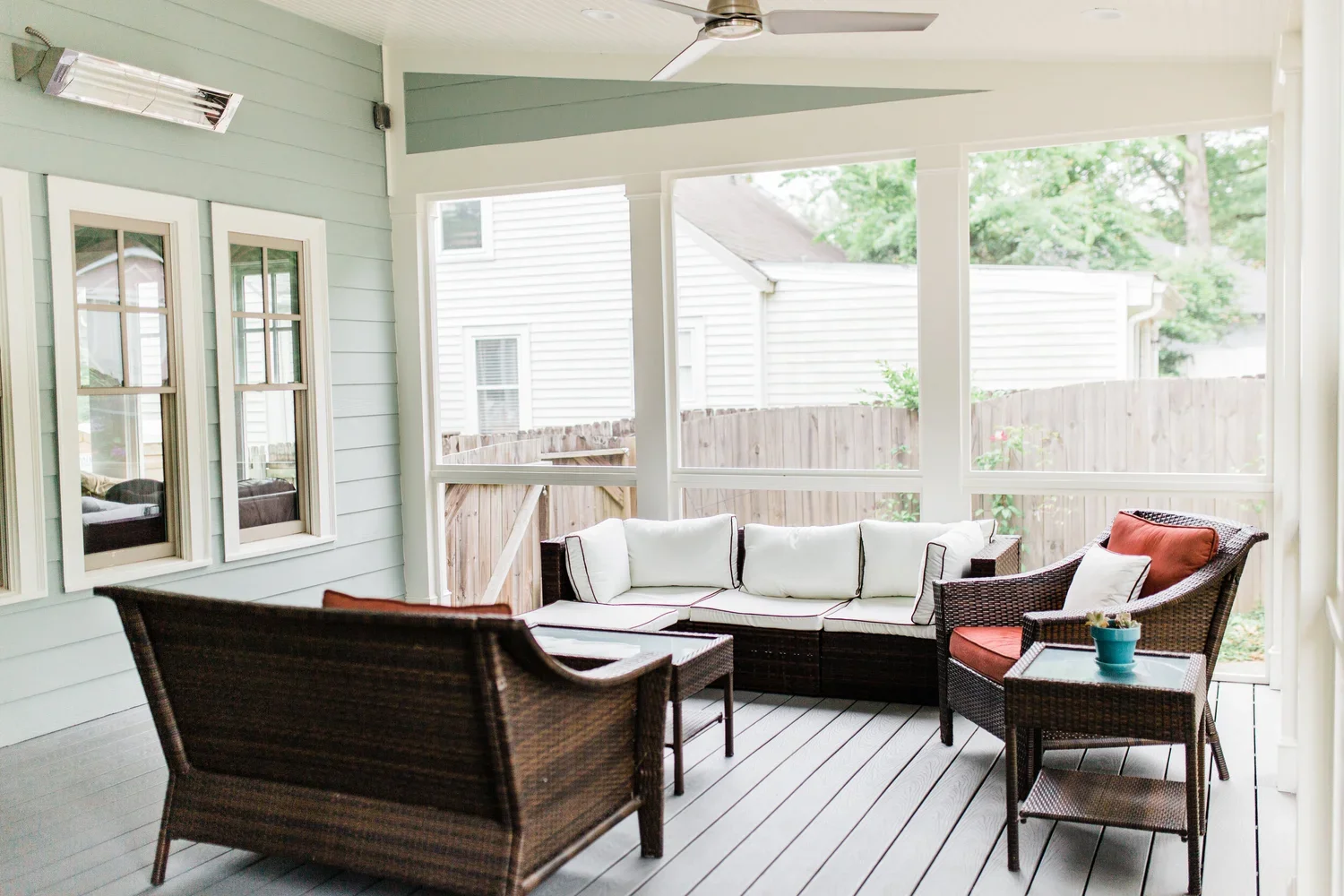 Covered outdoor porch with wicker furniture, white cushions, and small tables, surrounded by screened windows and a wooden fence.