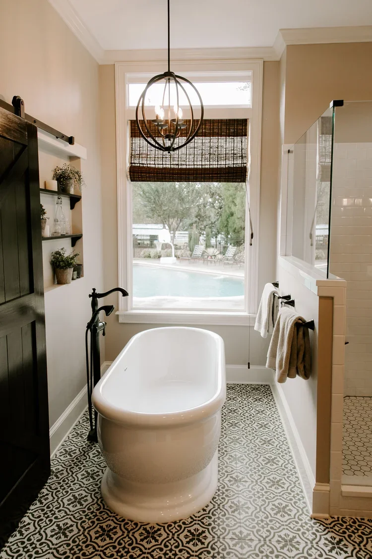 Freestanding white soaking tub, black-and-white patterned tile flooring, a dark wood sliding barn door, built-in wall shelving, frameless glass shower enclosure with white tile surround, oversized picture window, woven Roman shade, and matte black pl