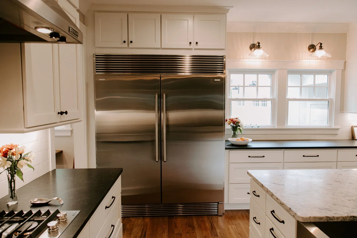 Modern kitchen with white cabinets, a large stainless steel refrigerator, black countertops, a stove, and two windows with flower arrangements.