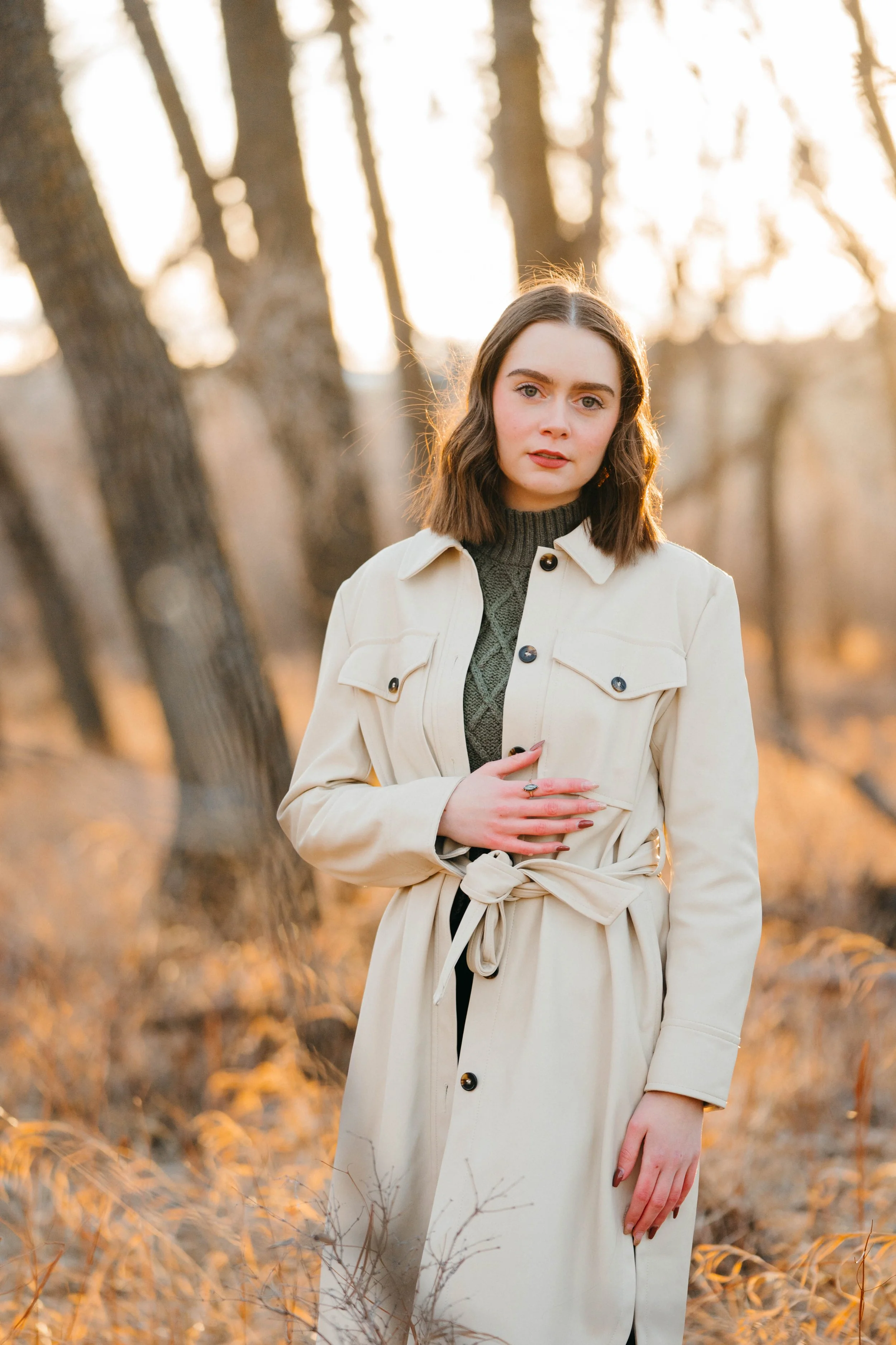 a photo of Myla wearing a long beige coat standing in front of some trees