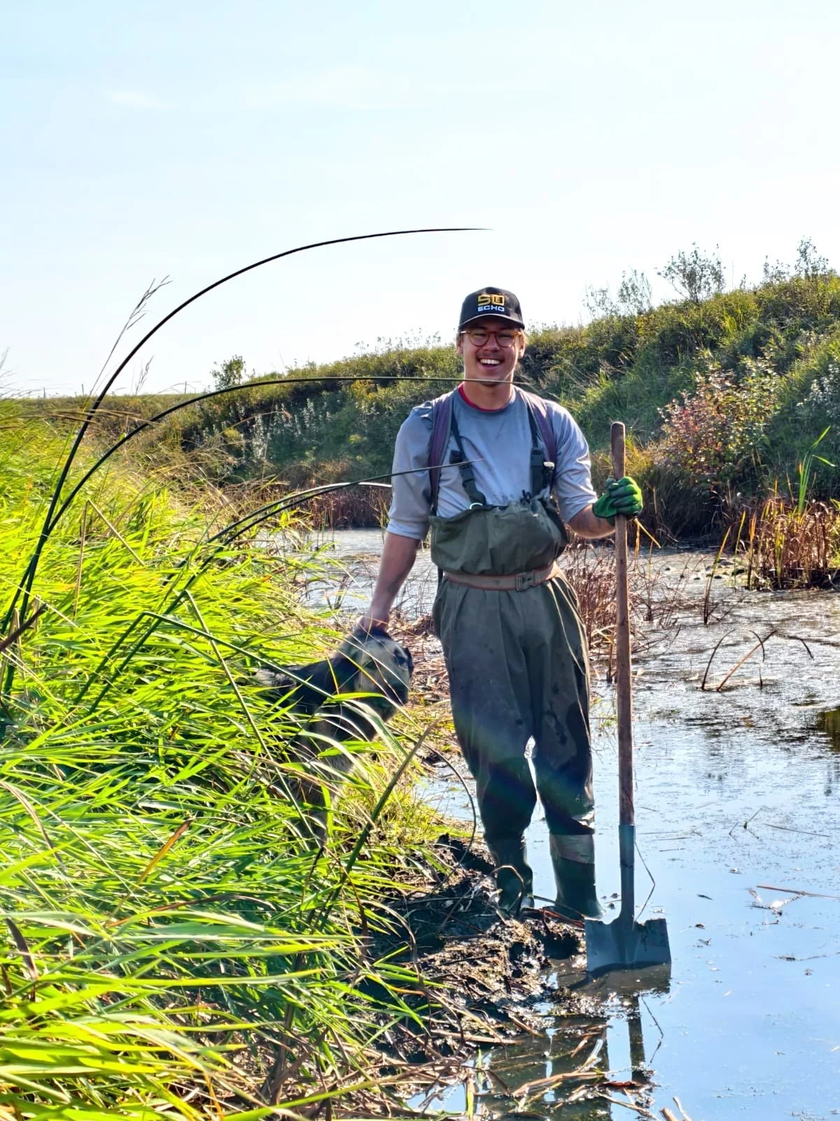 Elijah wearing waders and standing in mud while holding a shovel