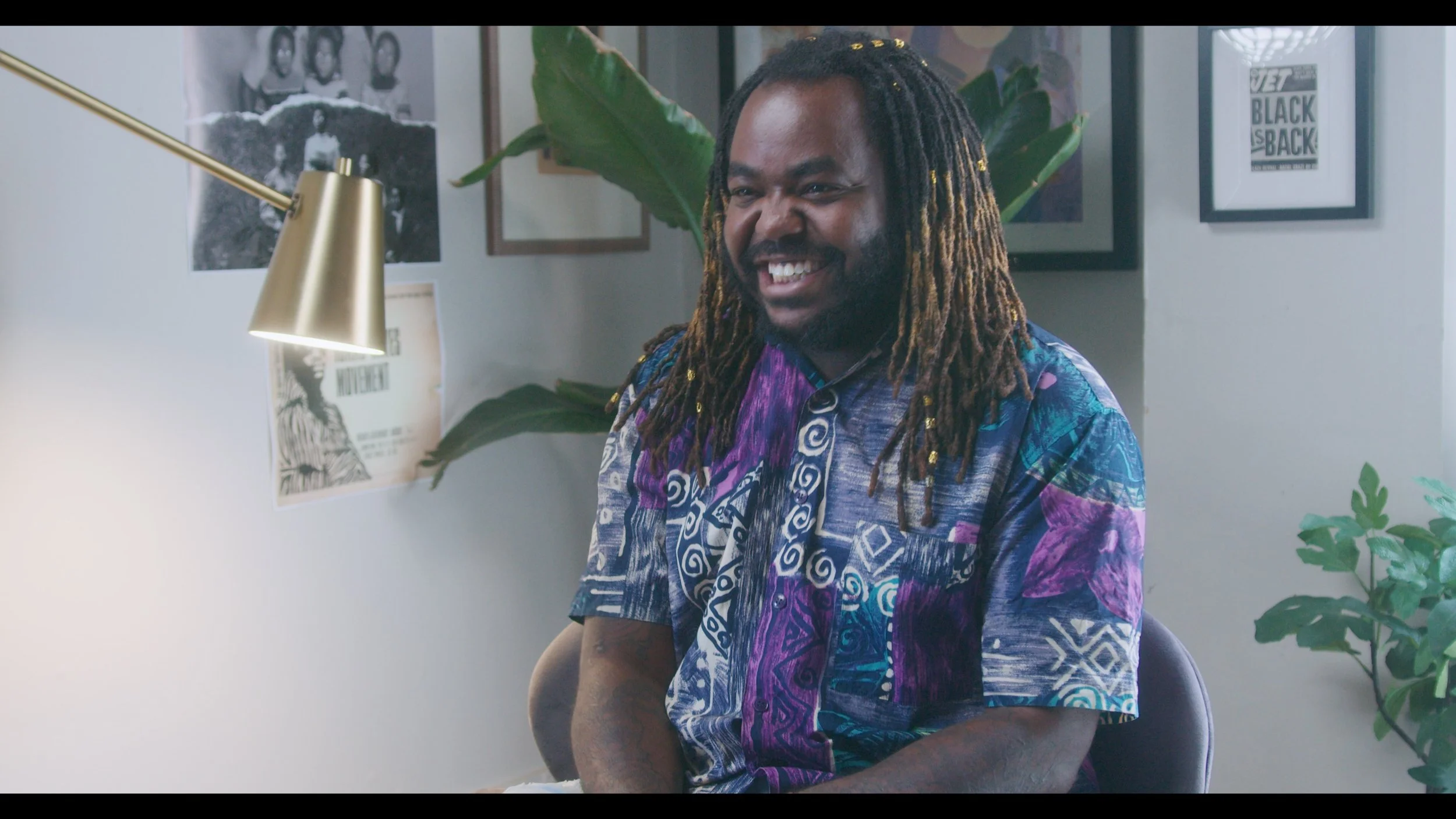 A man with dreadlocks smiling while sitting in a room decorated with framed pictures and posters on the wall, a large green plant, and a gold desk lamp.