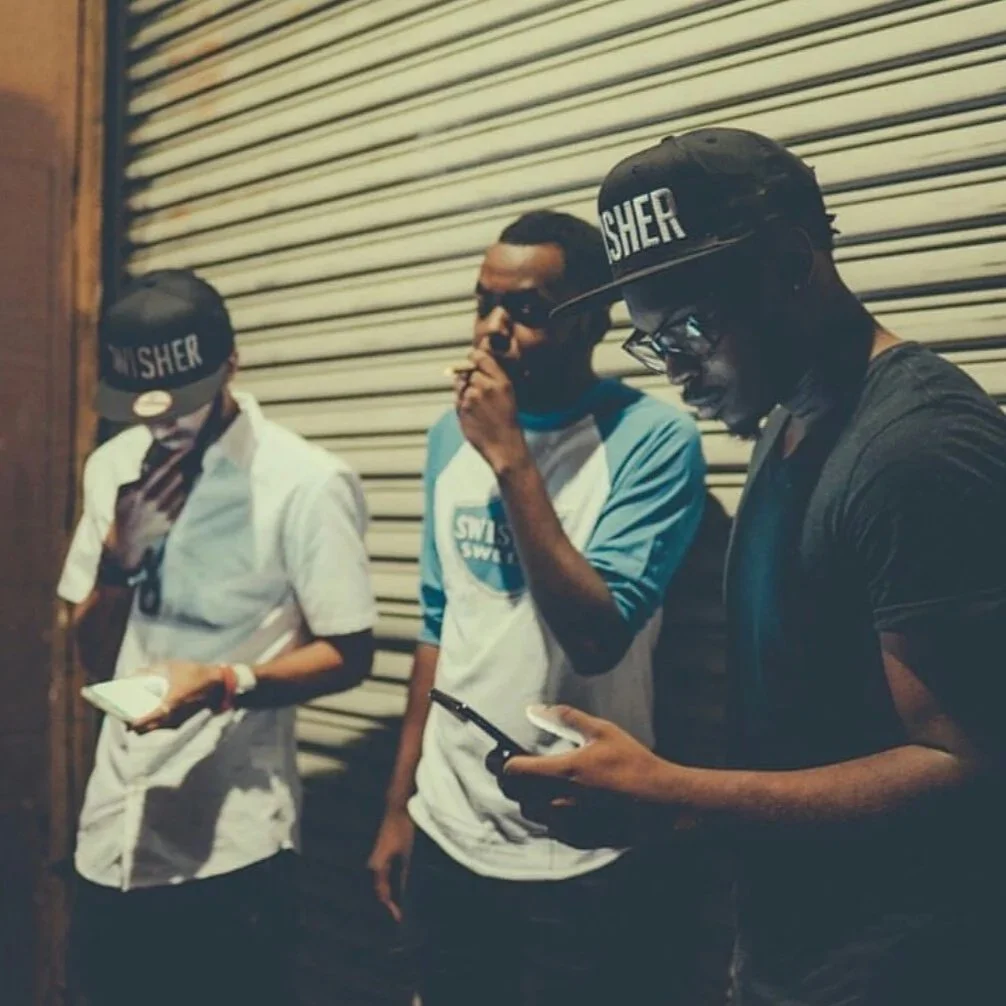 Three young men standing in front of a closed metal shutter at night, two are wearing hats with 'SHER' on them, one is using a smartphone, one is smoking a cigarette, and the other has a notebook.