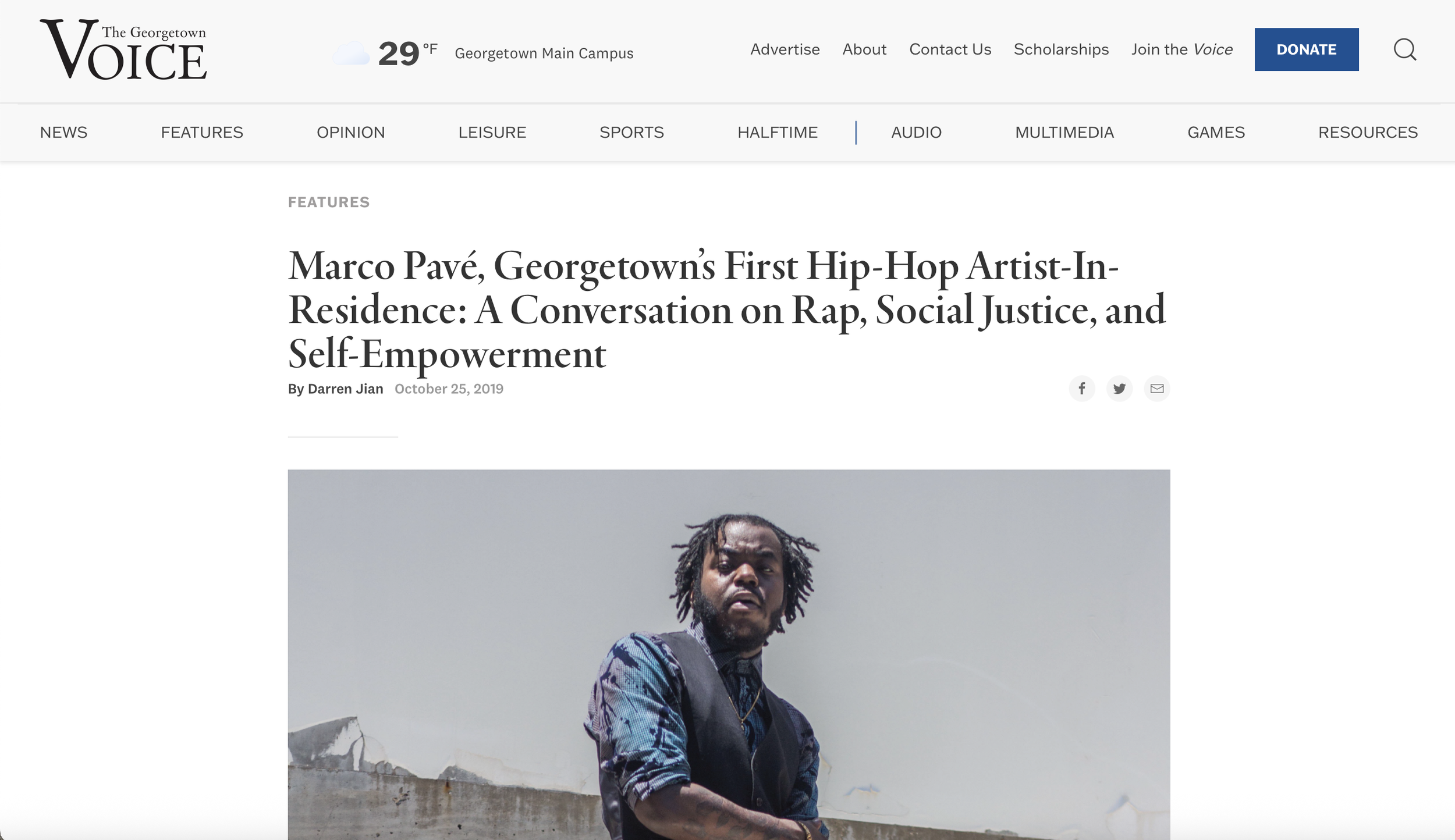 Portrait of a man with dreadlocks and a beard, wearing a patterned shirt and a backpack, standing outdoors against a concrete structure and clear sky.