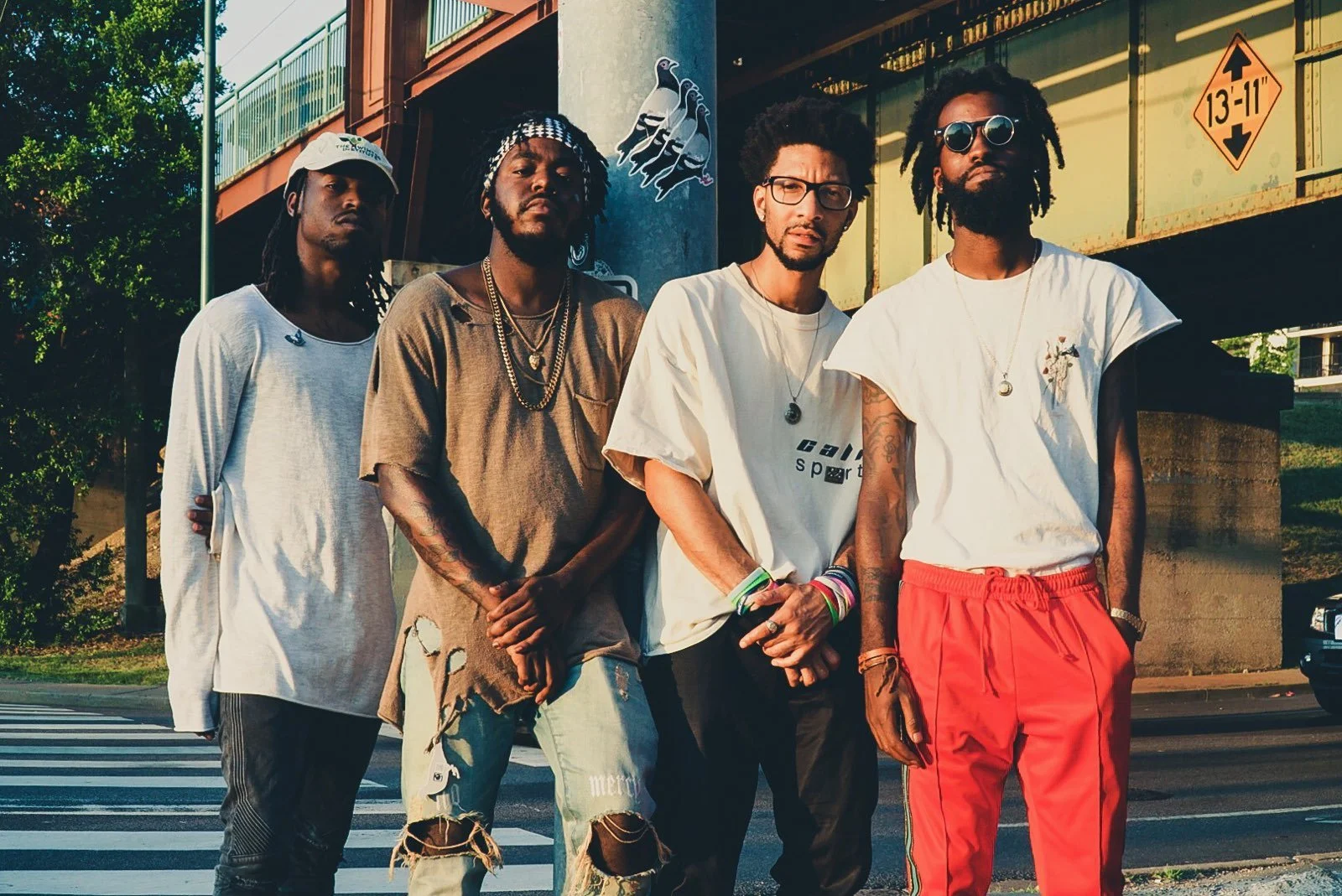 Four young men stand together on a city street at sunset, posing for a photo. They are dressed in casual, trendy clothing with accessories and tattoos, and are in front of an elevated train track and greenery.