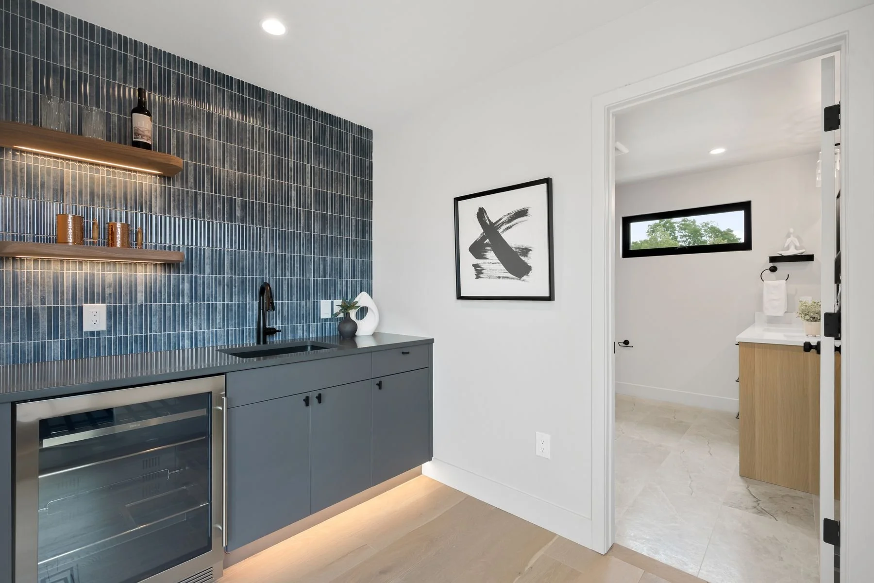 Modern kitchen with dark blue tiled wall, grey cabinets with built-in wine cooler, black faucet, and minimalist decor including a black and white abstract art piece.